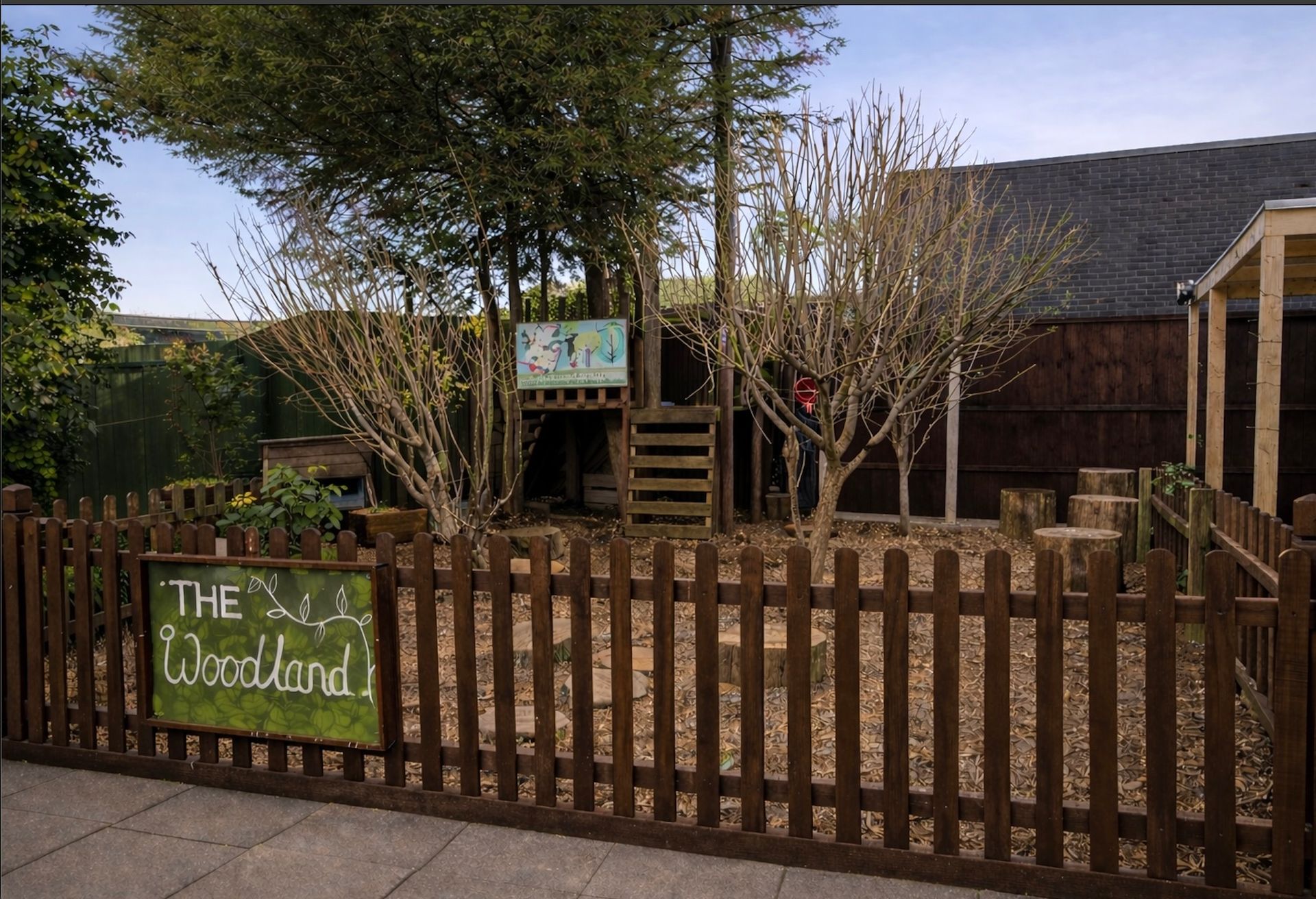 Wooden play area behind a fence with trees and a sign reading “The Woodlands”
