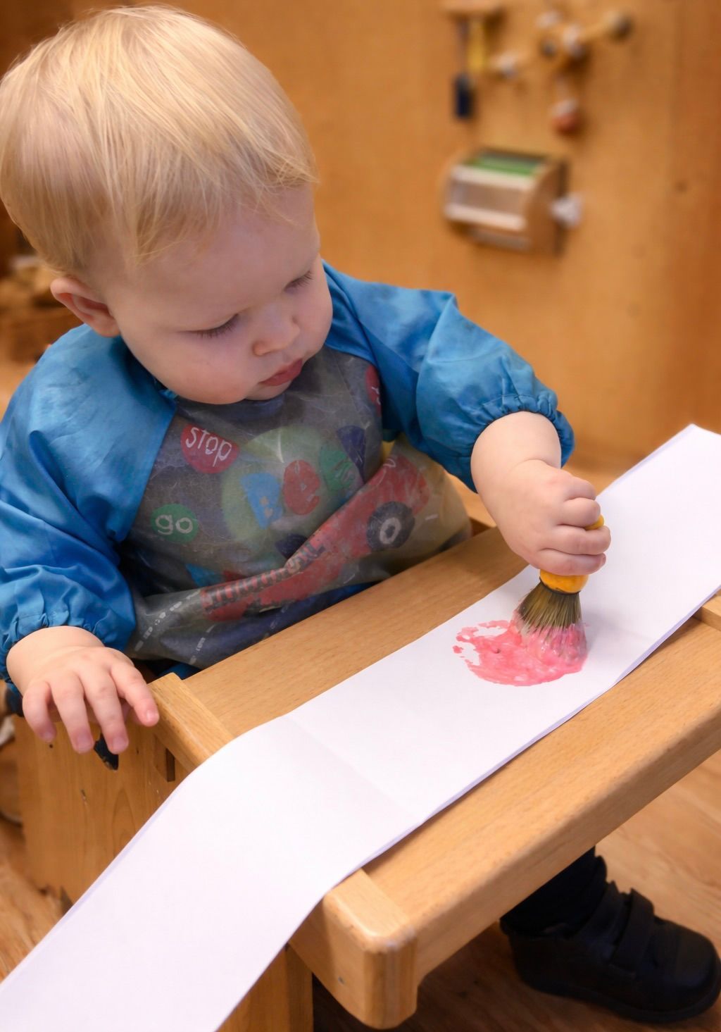 Toddler painting at a wooden table with red paint on a long white paper strip.