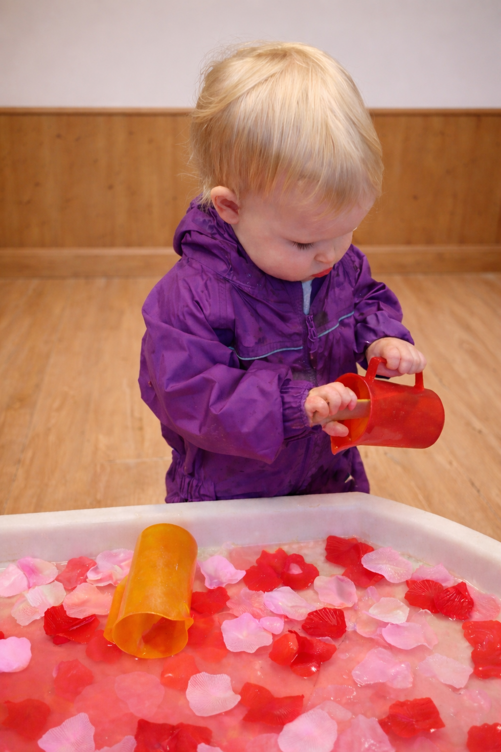 Toddler in a purple raincoat pouring red toy cups into a bin of pink water beads