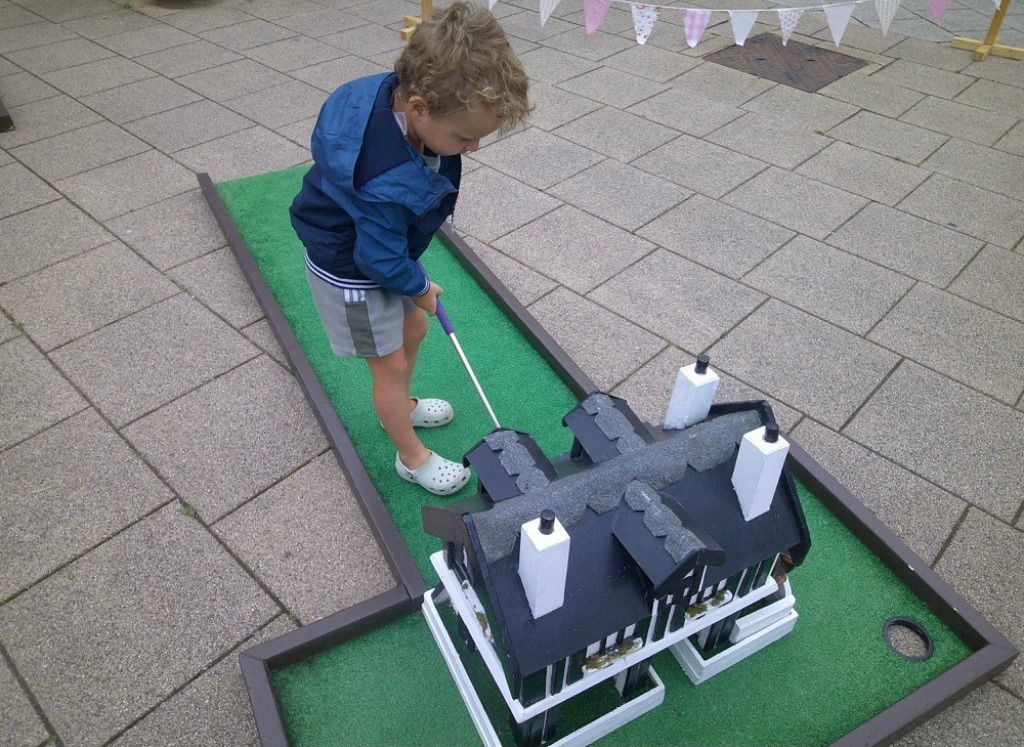 Child mini-golfing on a green putting lane beside a miniature castle model.