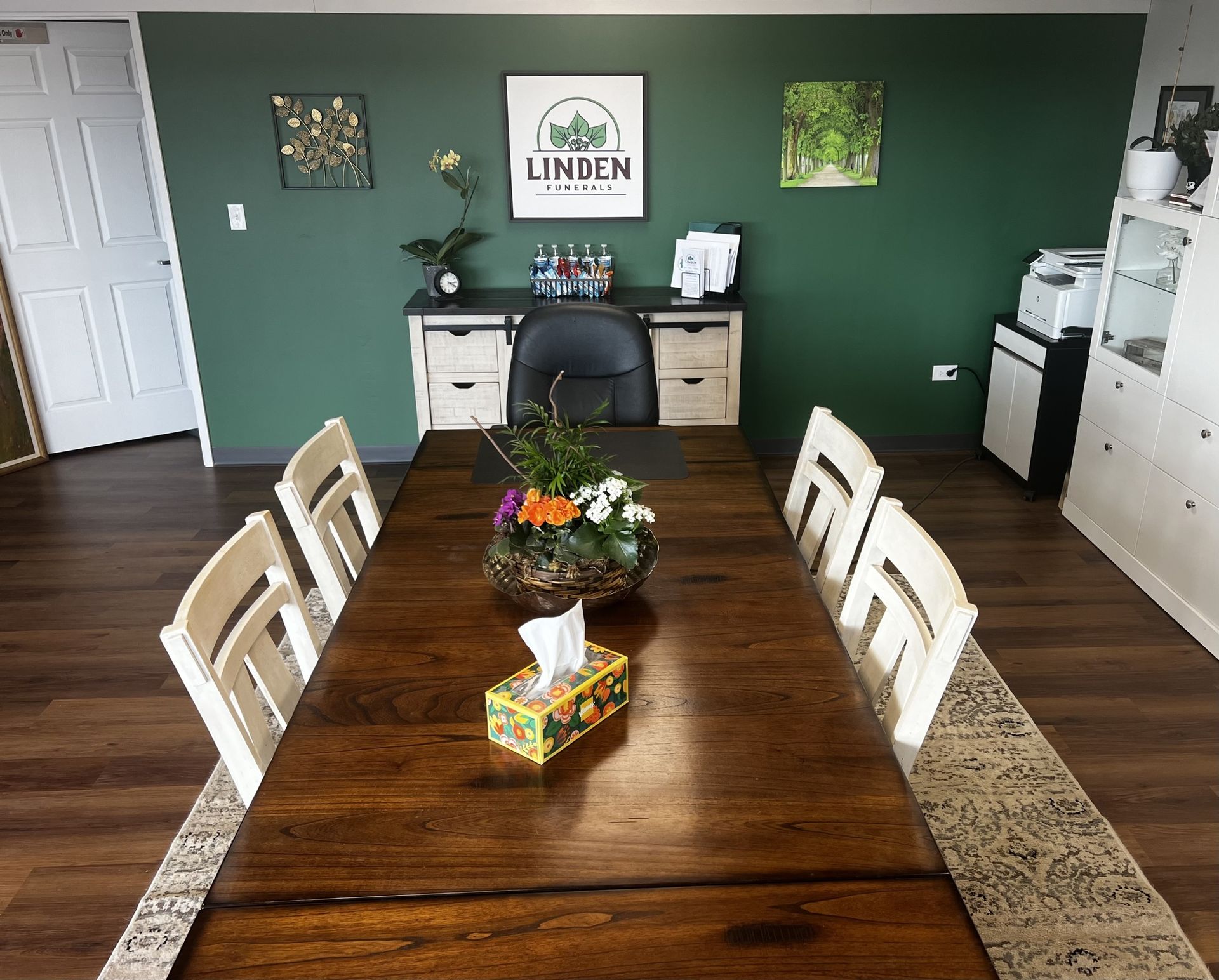 A conference room with a dark wooden table, white chairs, and a dark green wall featuring a desk and artwork.