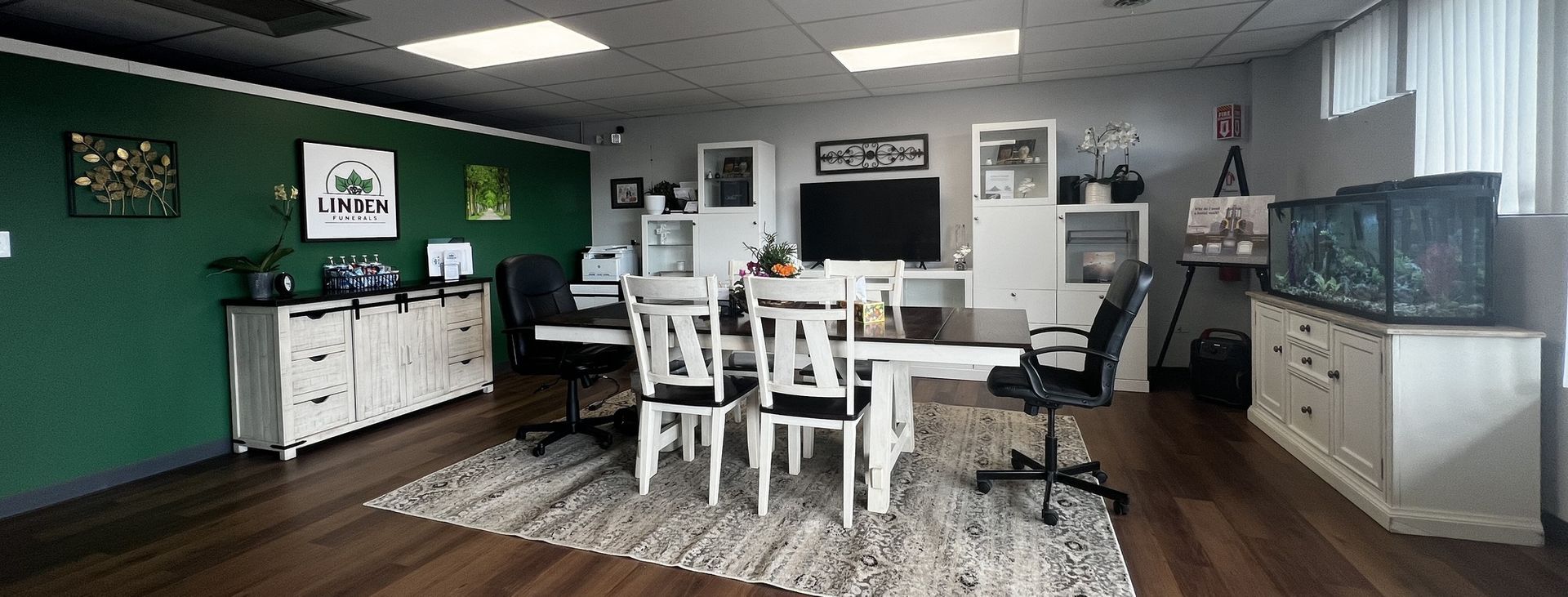 An office interior features a long white table with chairs, a green accent wall, and a large aquarium on a dresser.