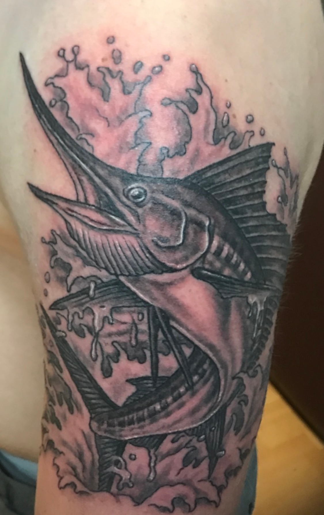 Black and grey marlin fish tattoo on an arm, with water splashes around it.