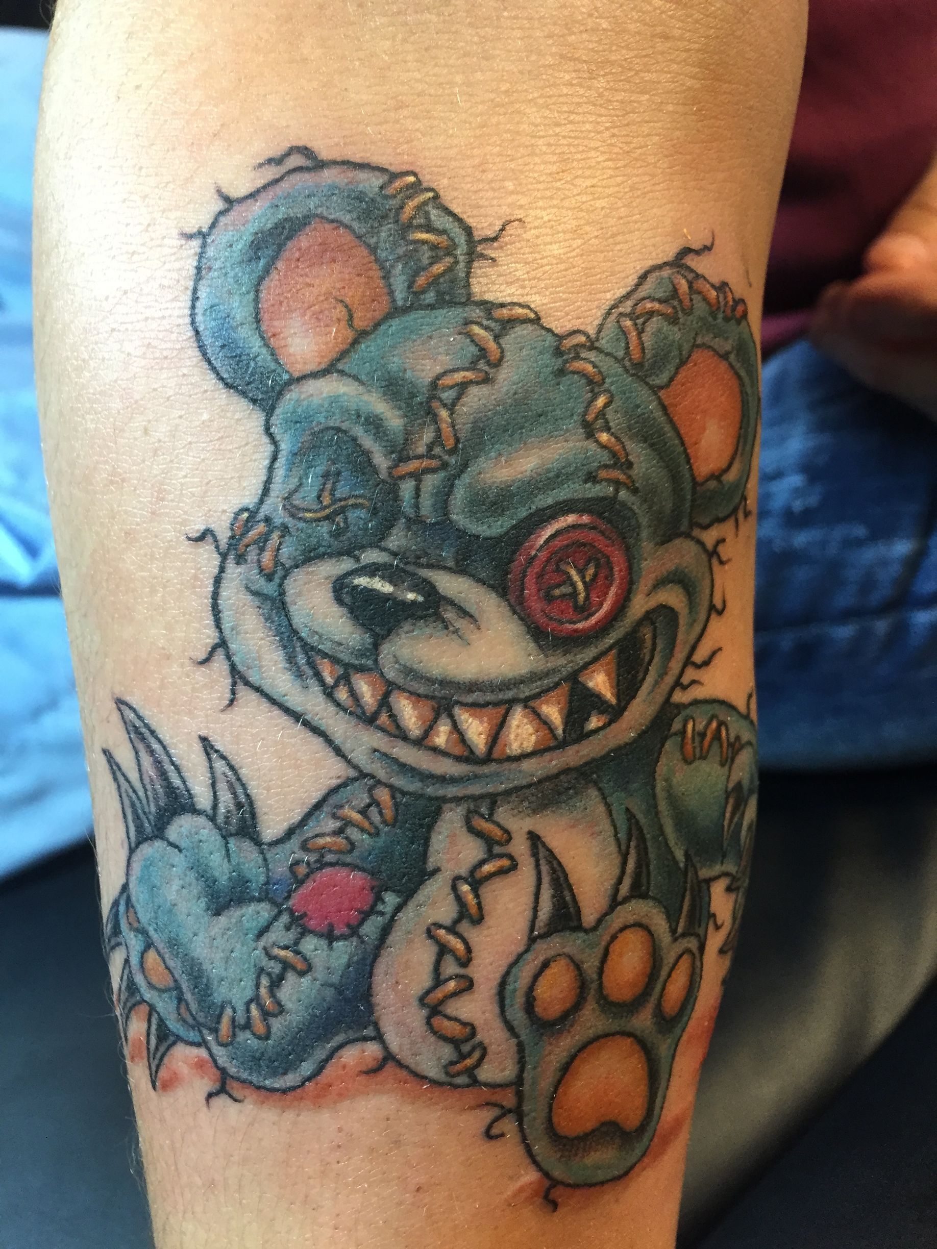 Tattoo of a stitched-up, grinning teddy bear with one button eye and sharp teeth.