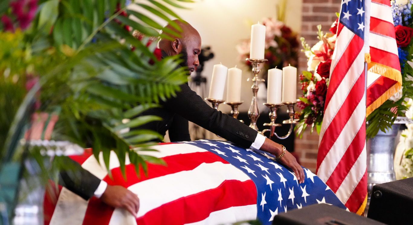 A person draped in a suit adjusts an American flag over a casket during a somber memorial service with lit candles.