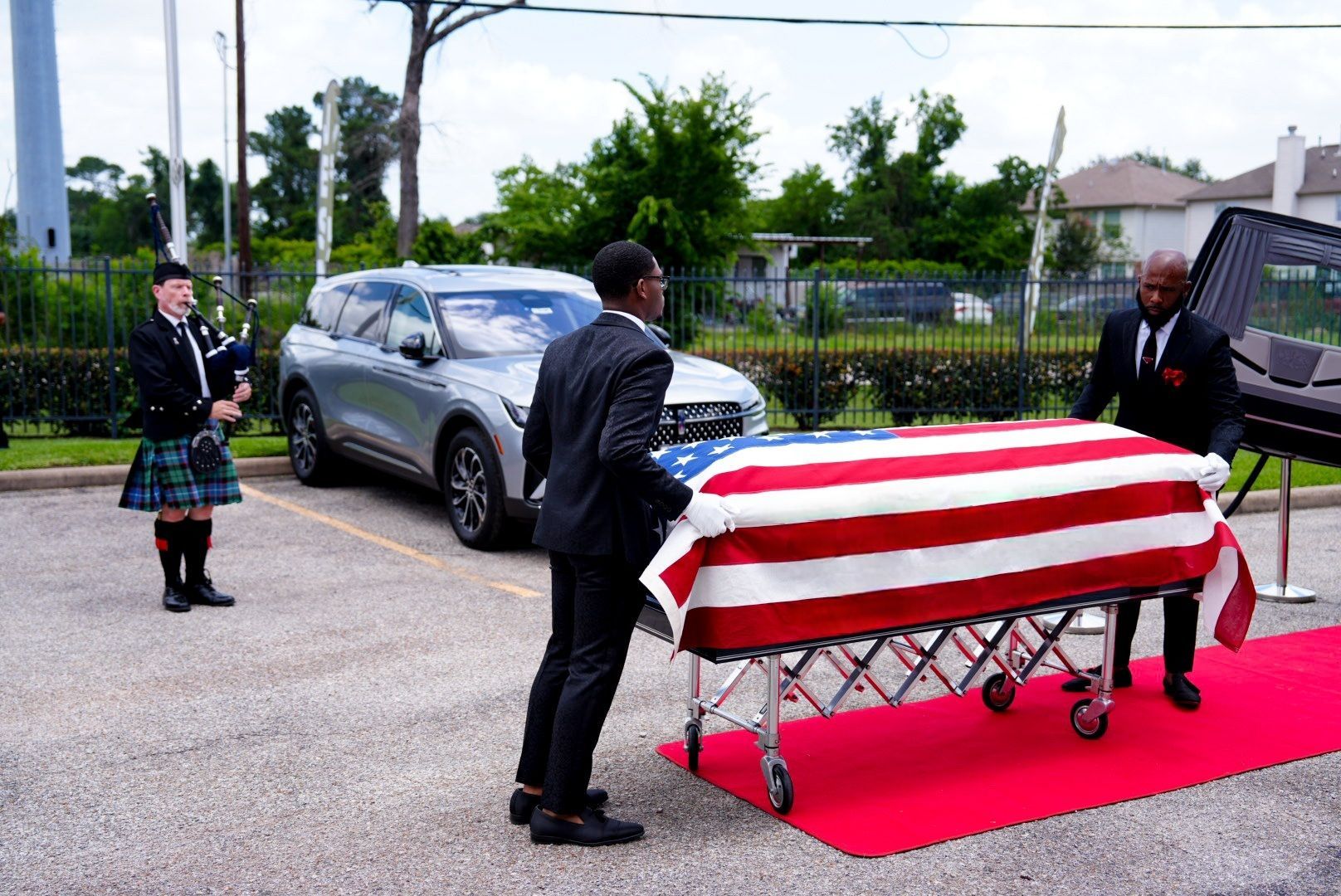 Two men in suits move a casket draped in an American flag, while a bagpiper in a kilt stands nearby in a parking lot.