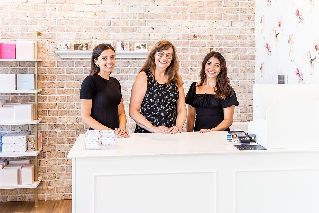 Three people stand behind a white counter in a boutique. Brick wall background, smiling, ready to assist customers.