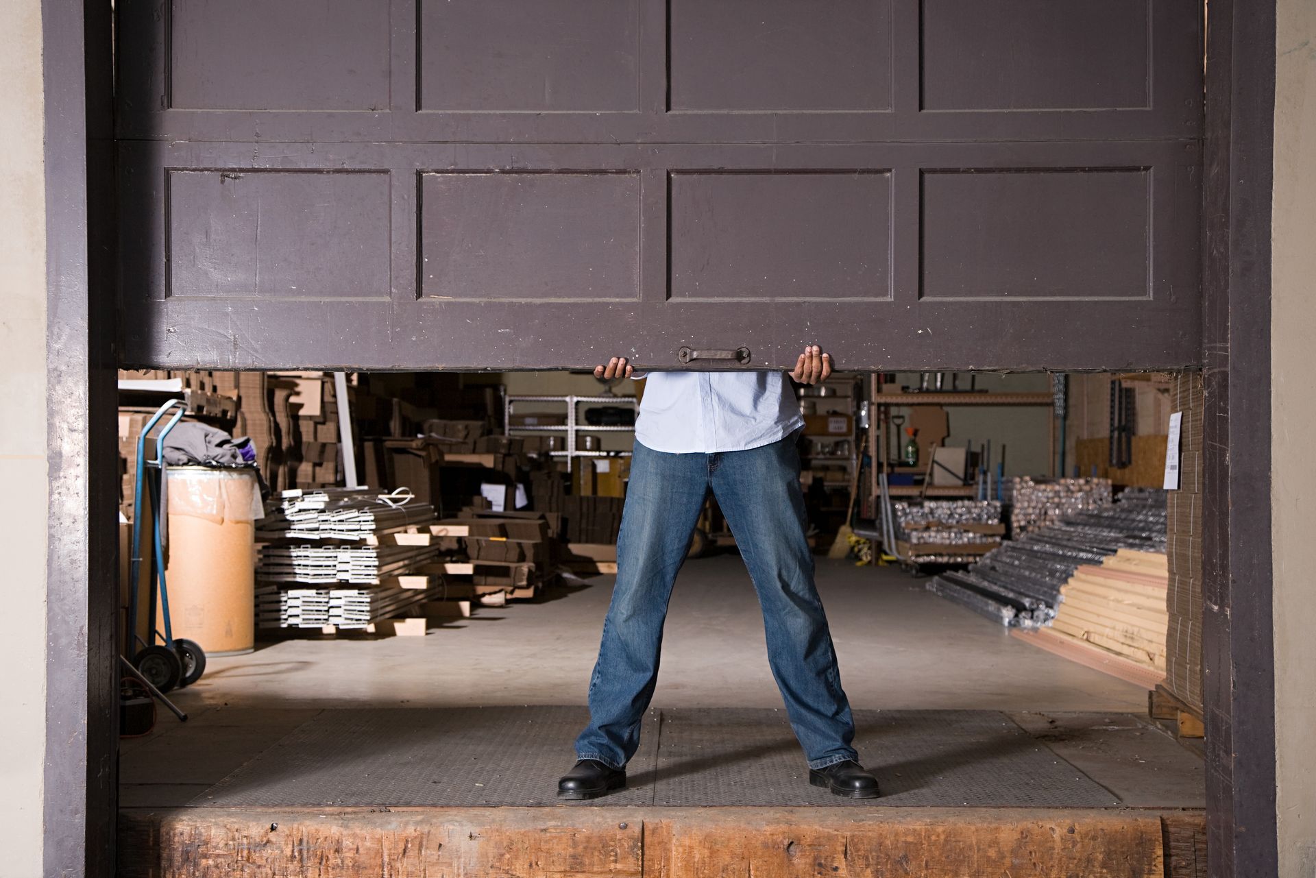 A person in a light blue shirt and jeans lifts a large, dark brown industrial garage door in a warehouse setting. A person in a light blue shirt and jeans lifts a large, dark brown industrial garage door in a warehouse setting.