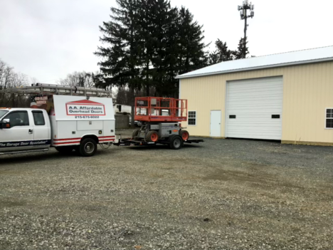 A man in blue overalls repairs a white garage door. A toolbox sits on the floor. A man in blue overalls repairs a white garage door. A toolbox sits on the floor.