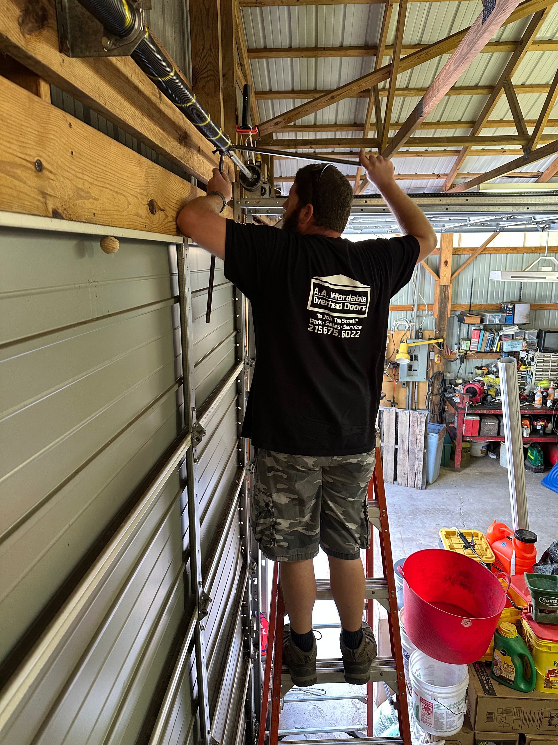 A worker wearing gloves uses a power drill to tighten a bolt on a garage door torsion spring assembly overhead.