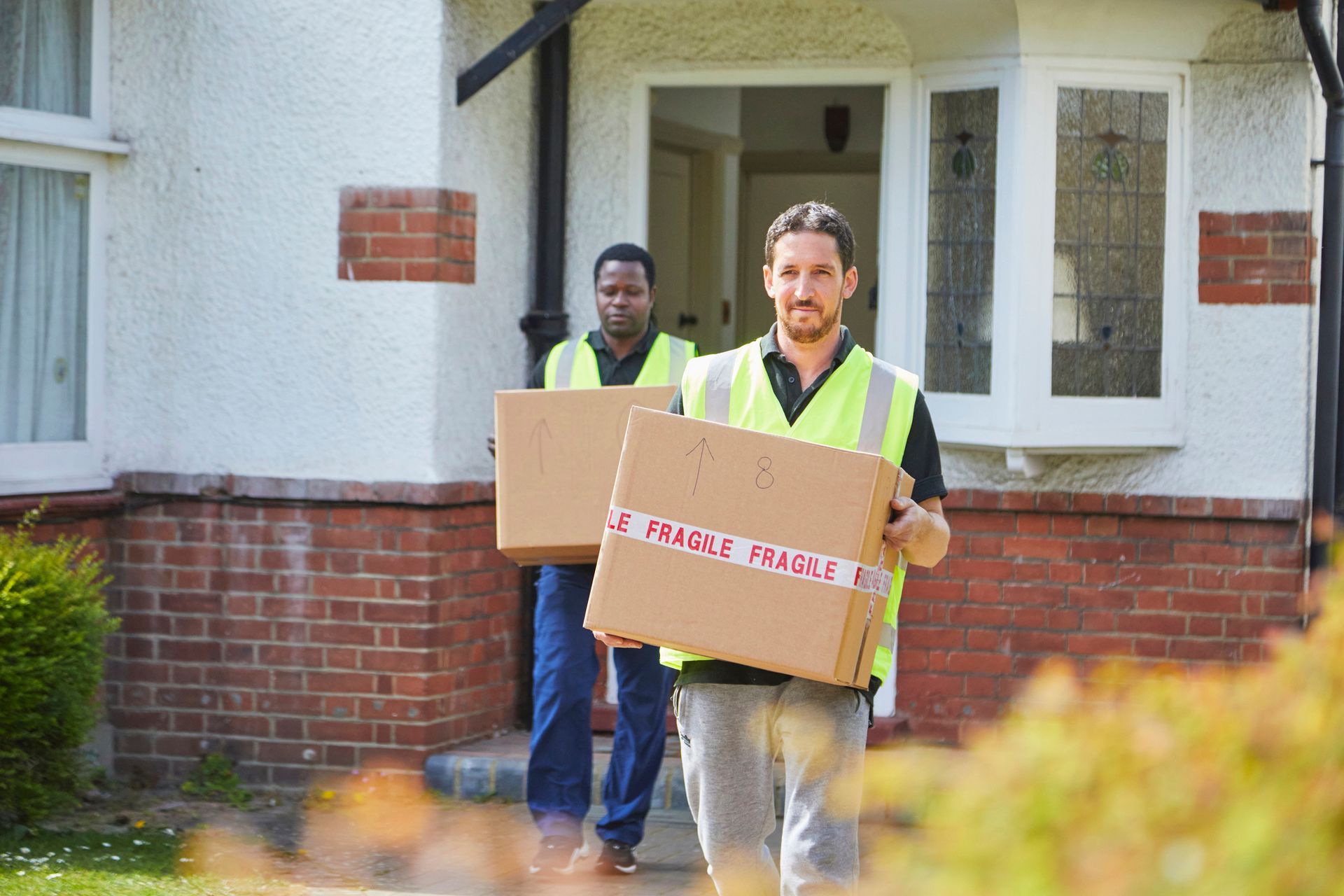 Two full service movers wearing reflective vests and carrying boxes, one of whom is smiling.