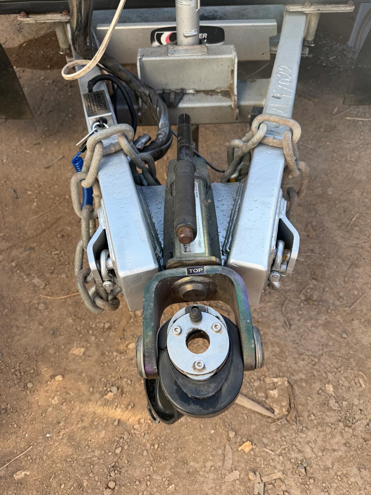 Close-up of a trailer hitch coupler and safety chains on a dirt ground.