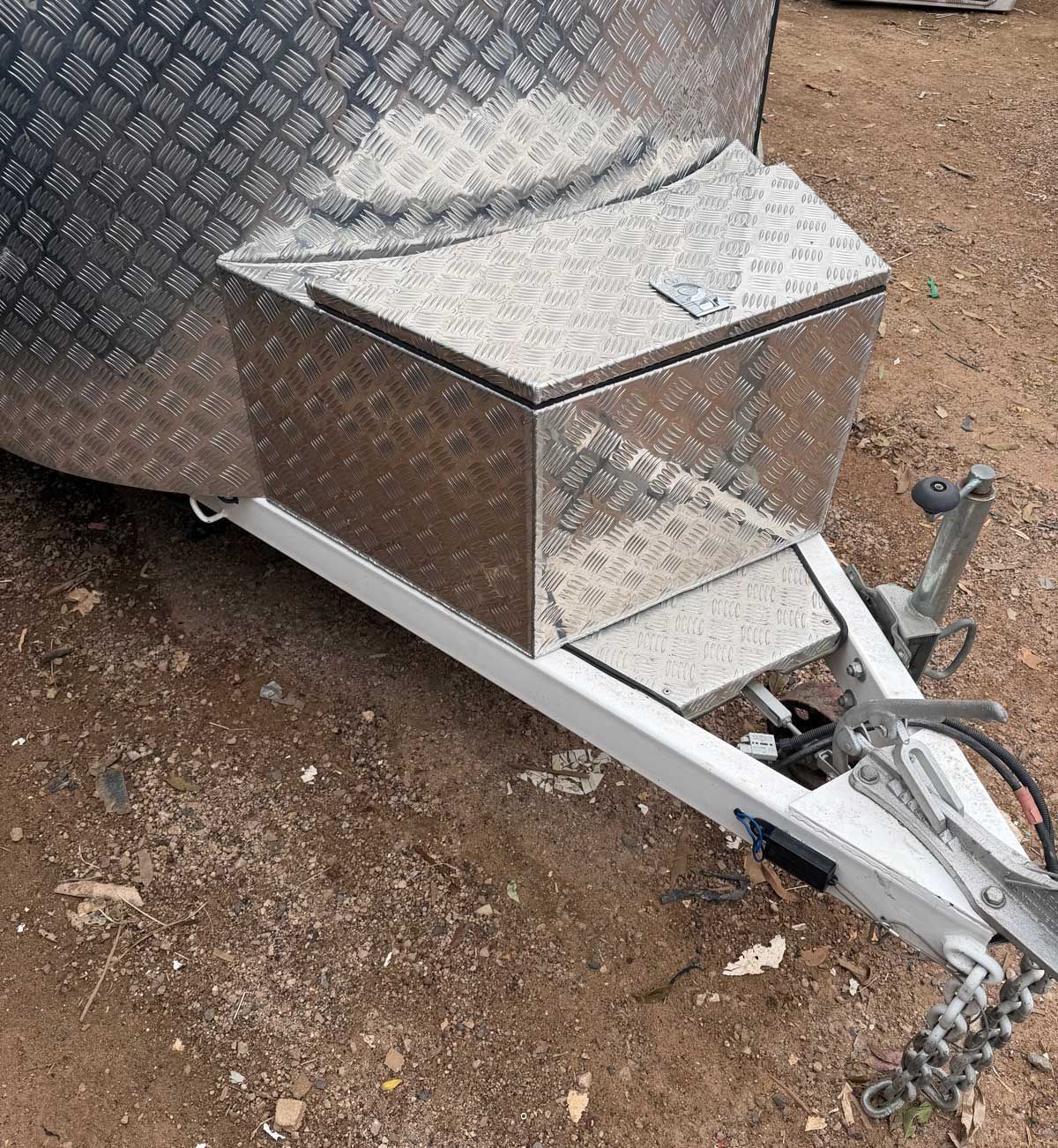 Silver diamond-plate trailer toolbox mounted on a utility trailer hitch, on gravel.
