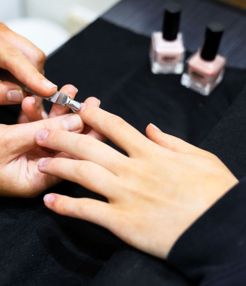 Manicurist trimming a client's fingernail with clippers; nail polish bottles in background.