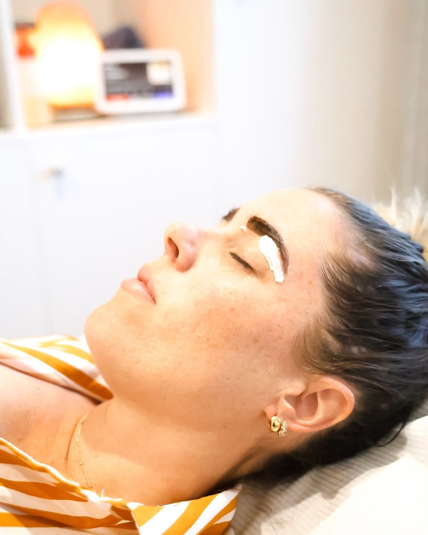 Woman with cream on her eyebrow, eyes closed, resting on a white surface.