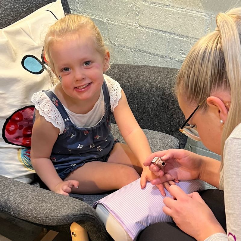 Young child smiling while getting a manicure; adult is painting nails indoors.