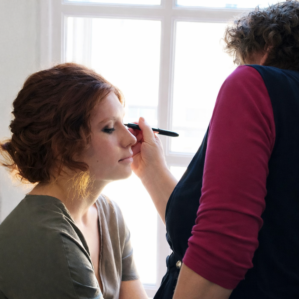 Woman getting makeup applied next to a window. Makeup artist in burgundy and black shirt.