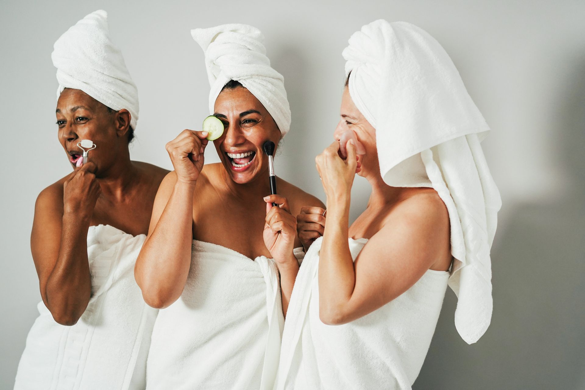 Three women wrapped in white towels, laughing while applying skincare in front of a gray wall.