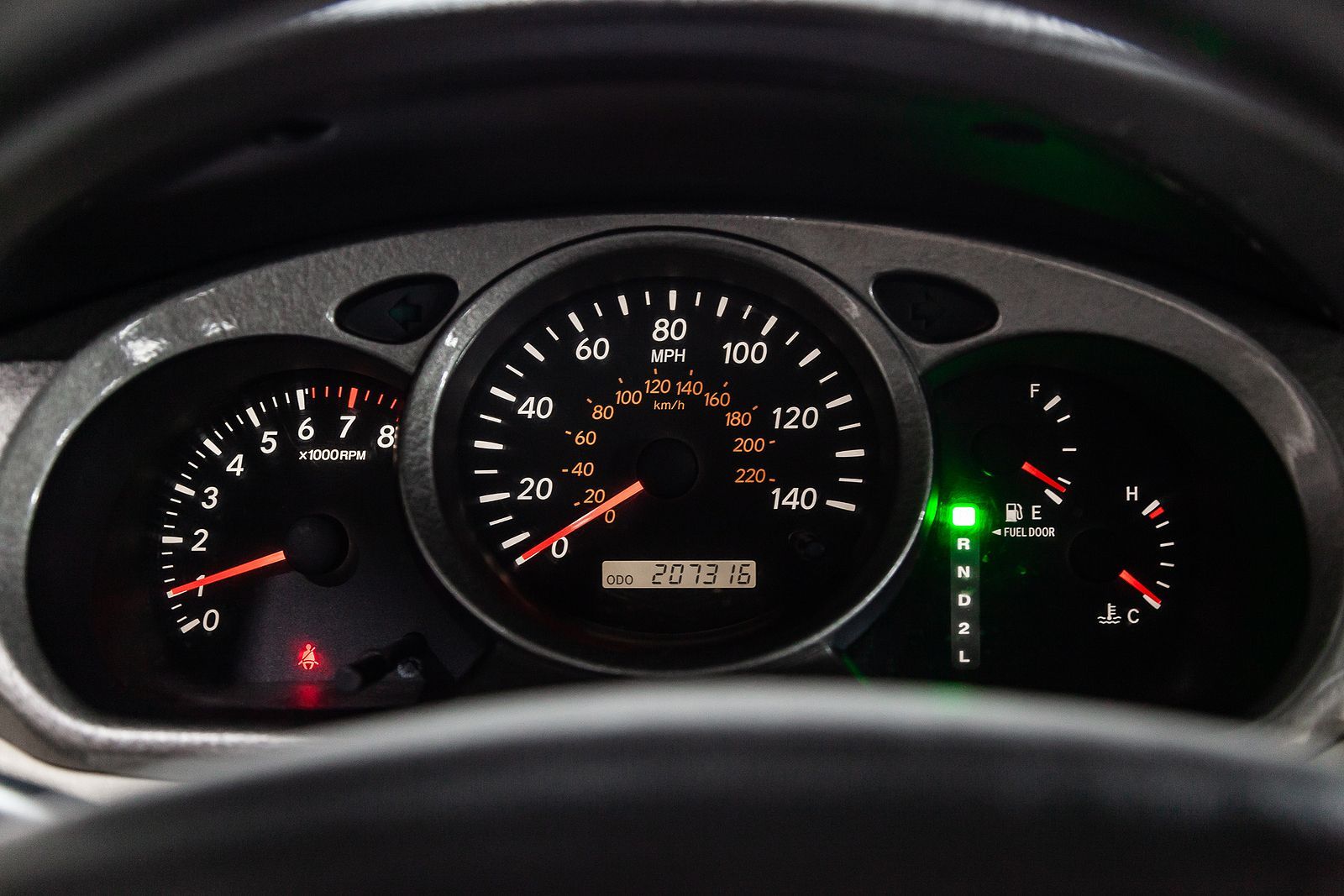Car dashboard with gauges: speedometer, tachometer, and fuel gauge, illuminated with white and orange lights.