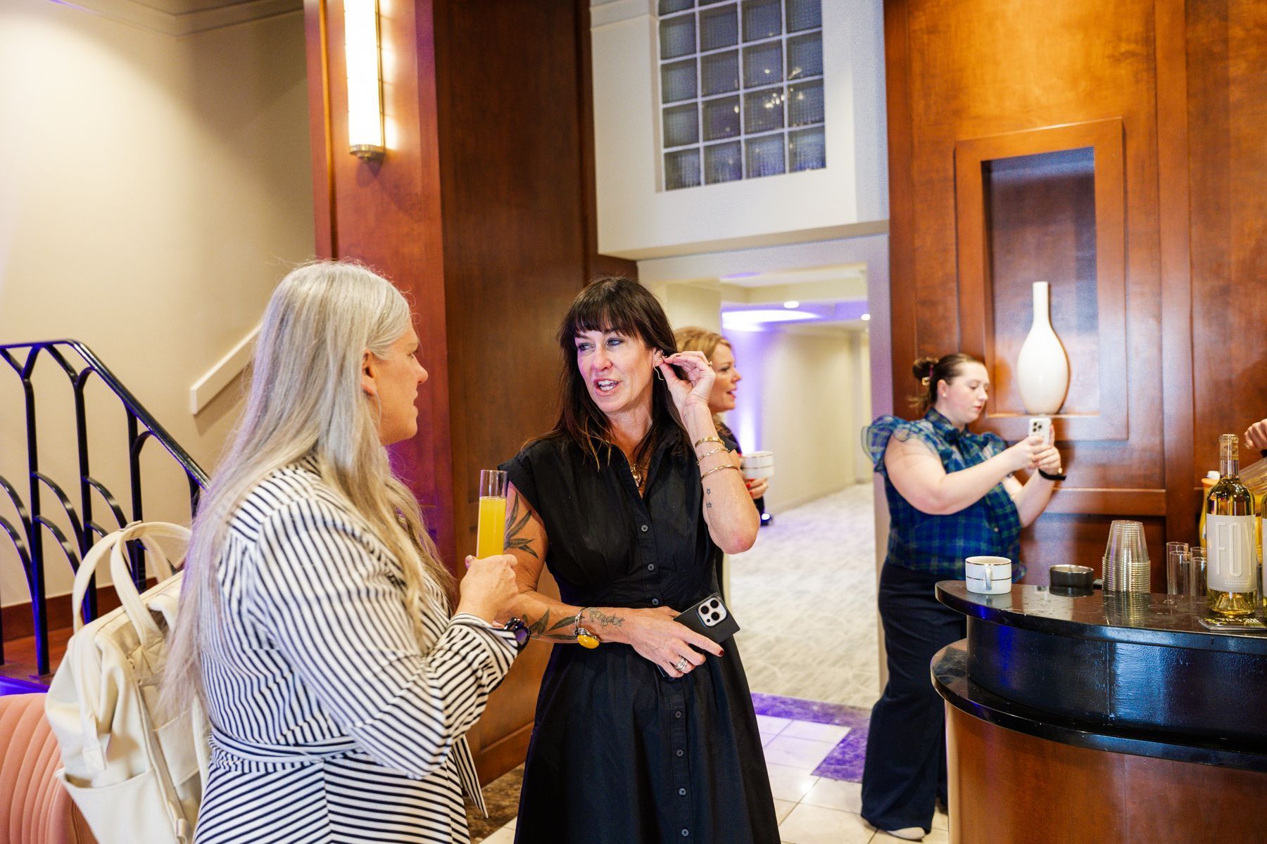 People at an event. A woman in a black dress adjusts her earring, holding a drink. Another woman in stripes, and another taking a photo.