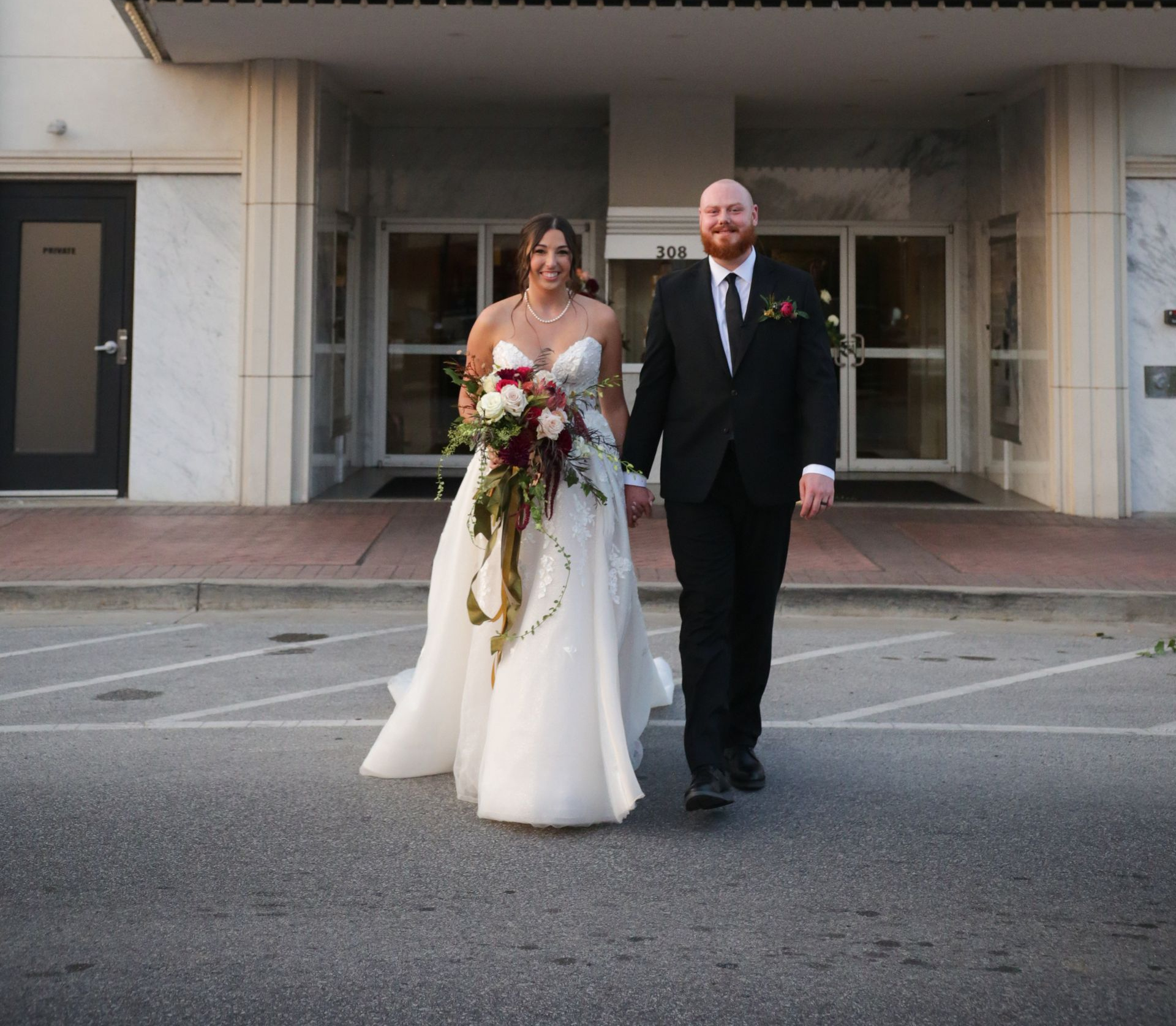 Bride and groom holding hands, exiting a building, wearing wedding attire.