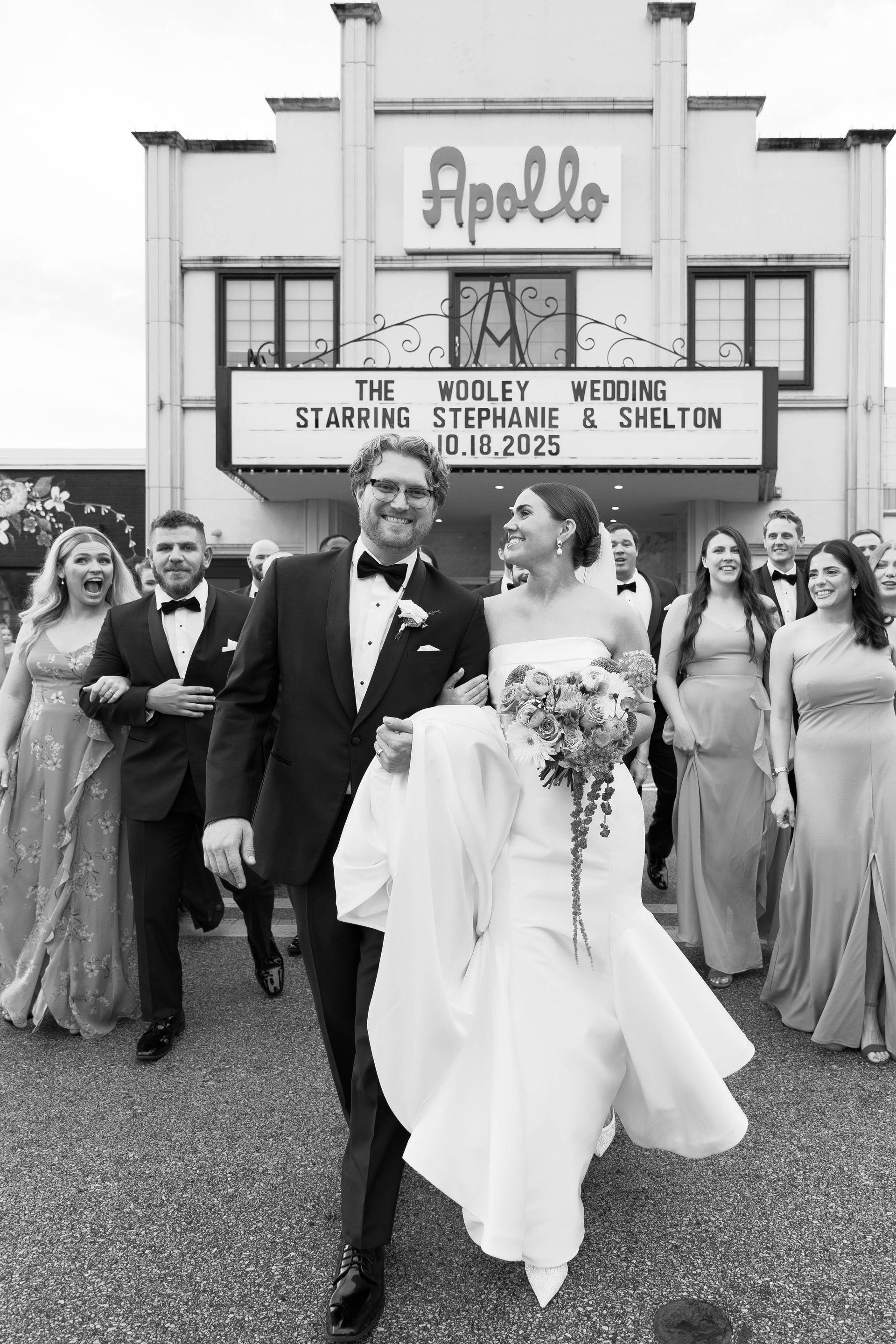 Wedding party exits Apollo Theater. Bride and groom smiling, holding hands. Bridesmaids and groomsmen follow. Black and white.