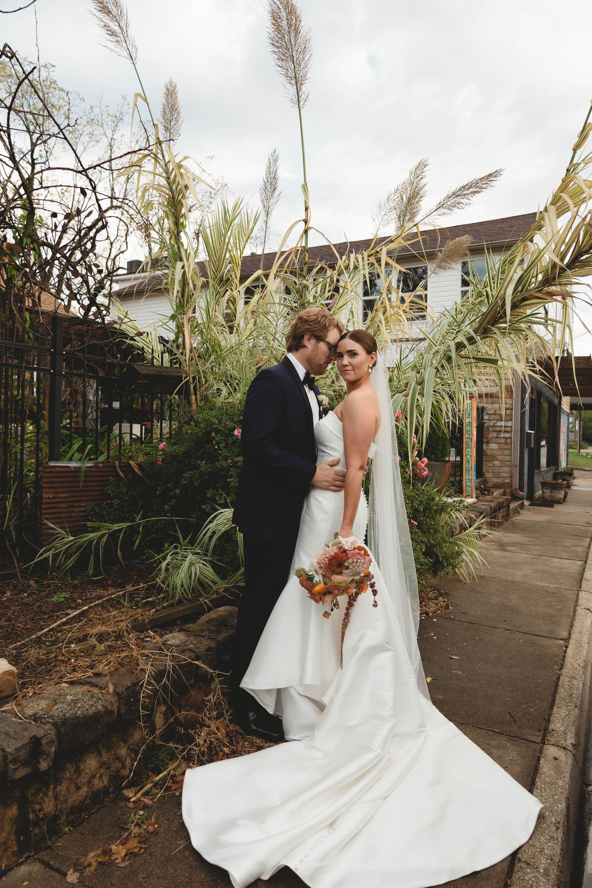 Bride and groom embrace outdoors, near a building with foliage. The bride wears a white dress; the groom, a dark suit.
