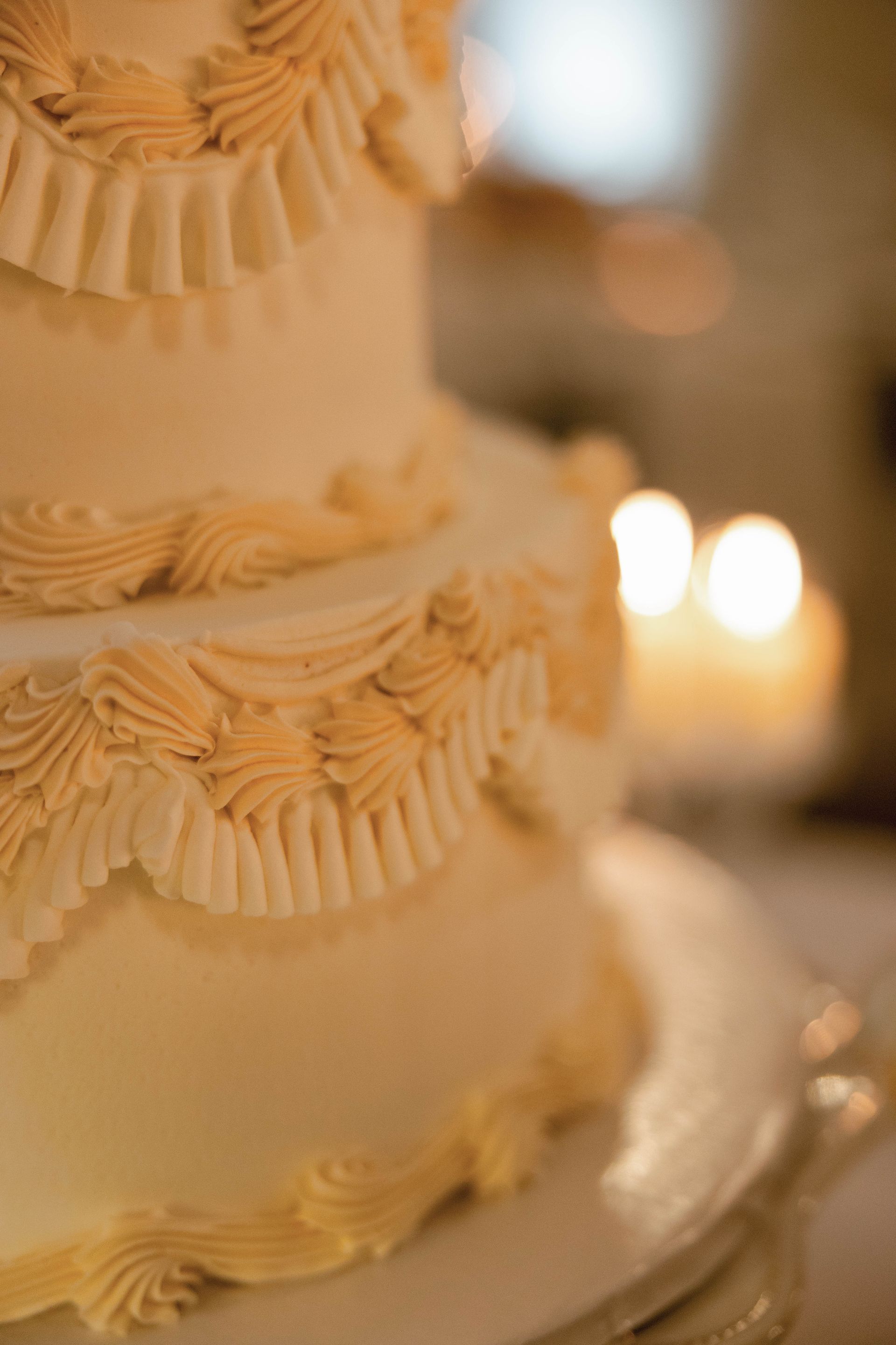 Tiered wedding cake with beige frosting and decorative swirls, candles in background.