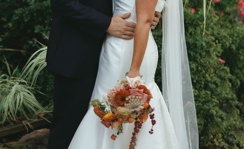 Bride and groom embrace outdoors, bride in white dress holding bouquet.