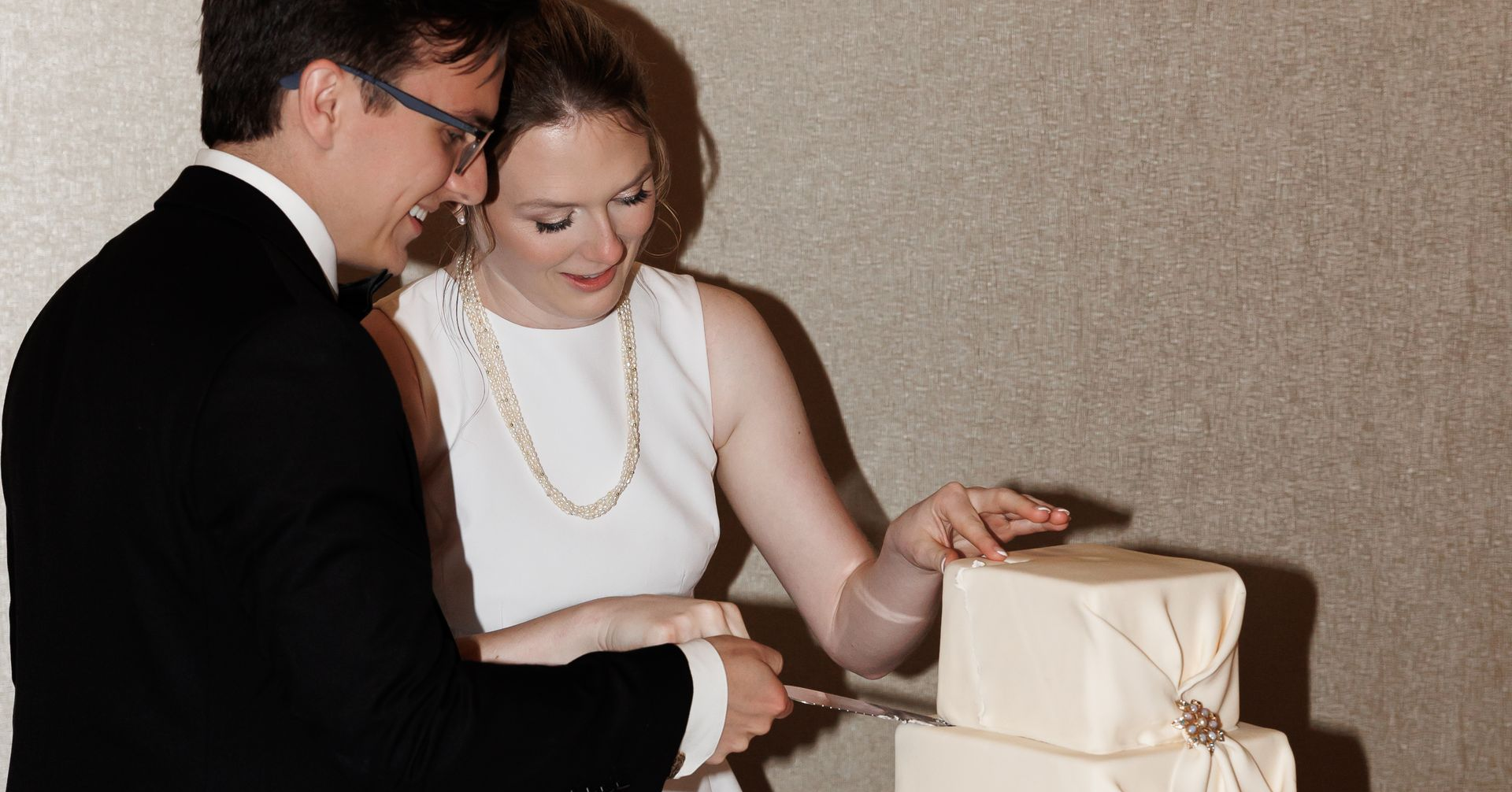 Couple cutting a three-tiered wedding cake. The bride wears a white dress, a necklace. The groom wears a black suit, glasses.