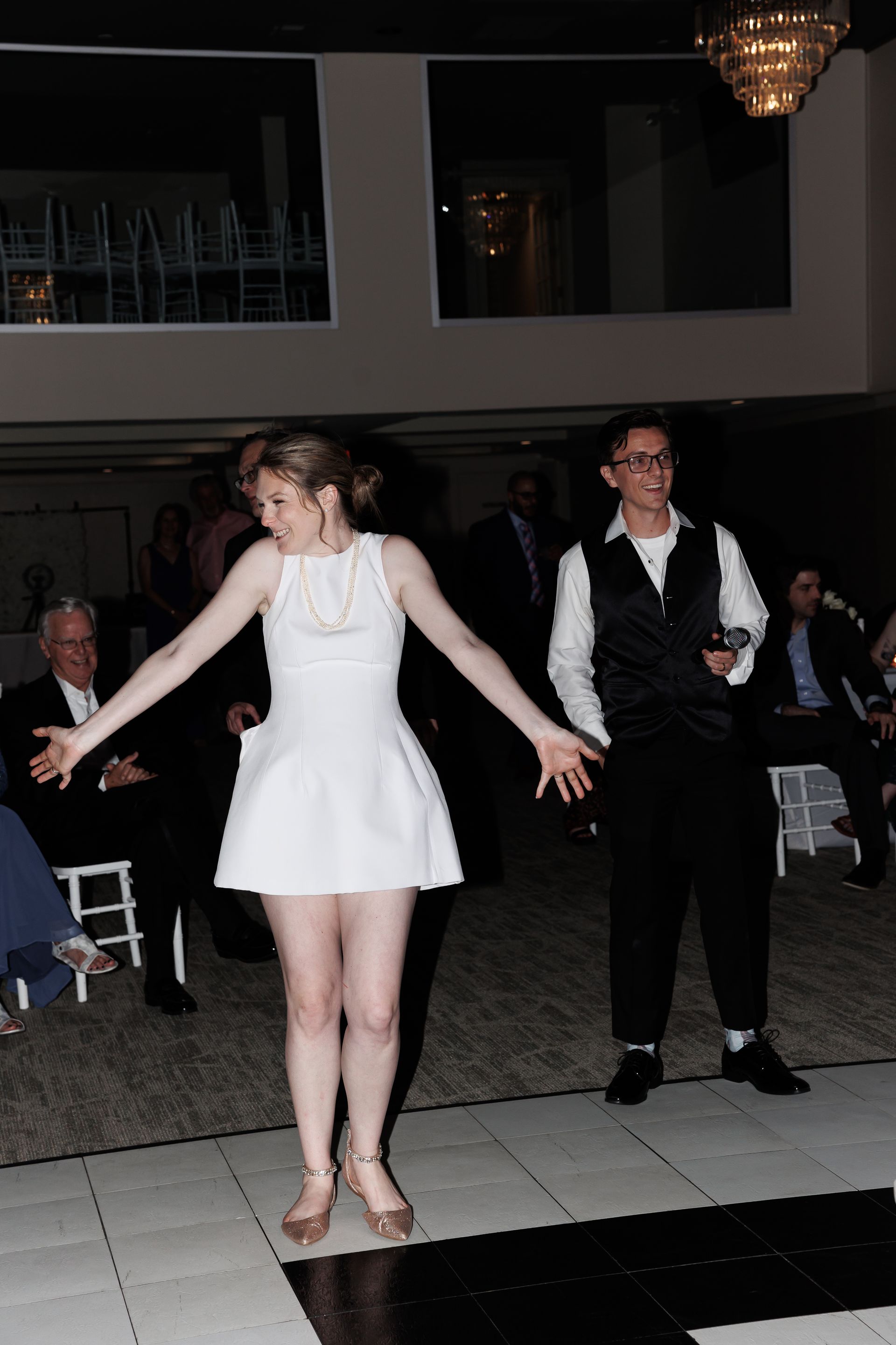 Woman in white dress dances with man in vest at a party; black and white dance floor.
