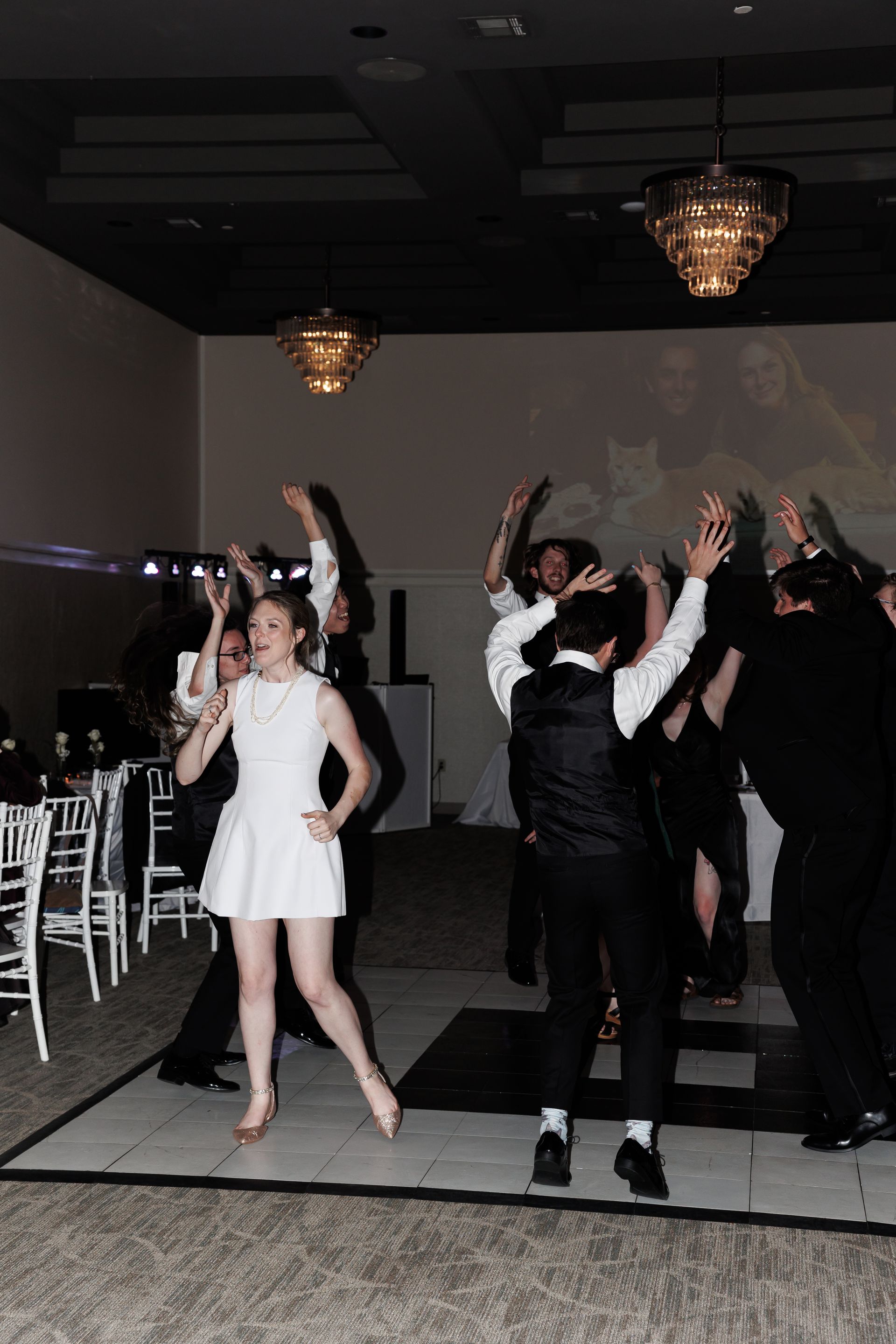 People dancing at an event, wearing formal attire. White dress, dark vests, chandelier lighting.