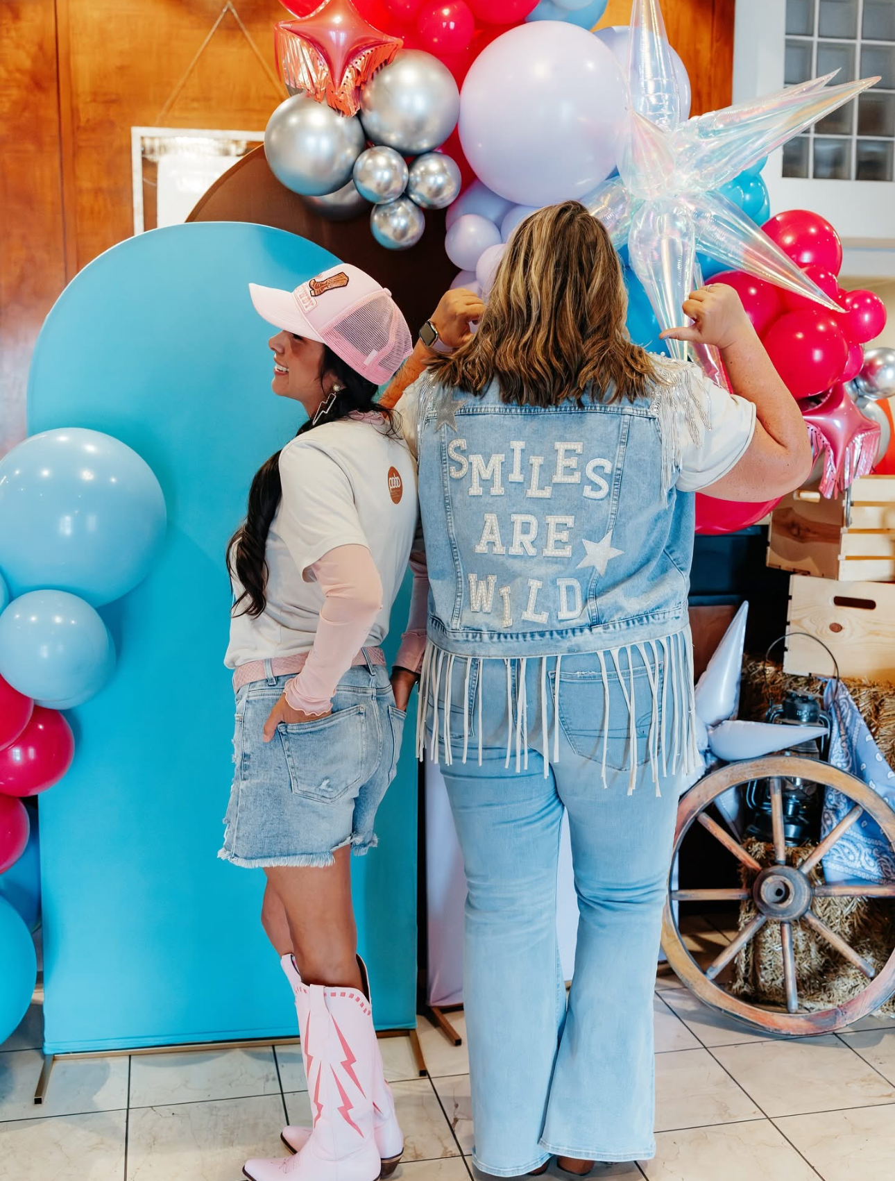 Two people posing in front of a balloon arch, wearing denim and pink cowboy boots; “Smiles are Wild” on denim vest.