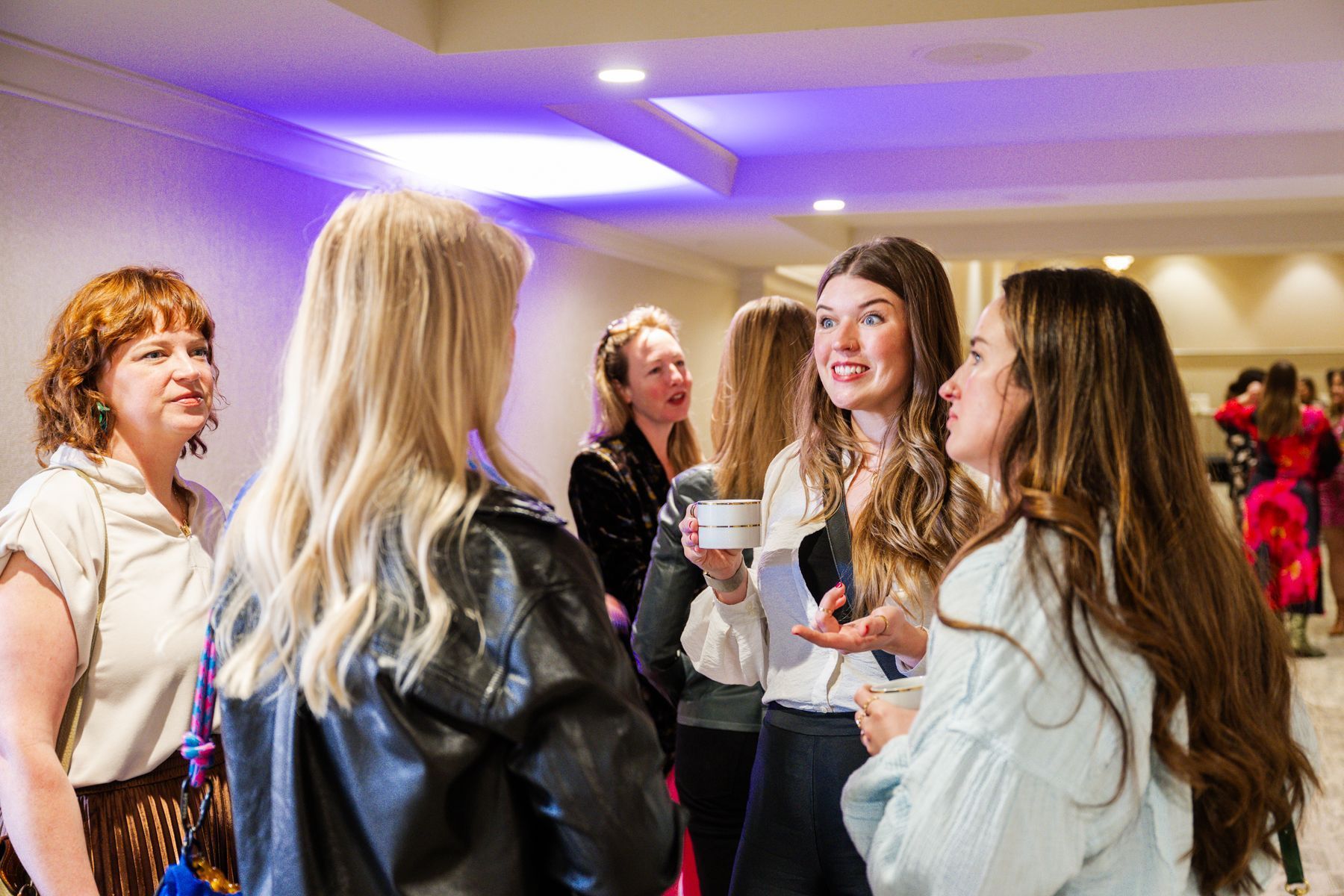 A group of women conversing in a brightly lit hallway; one holds a cup and papers.