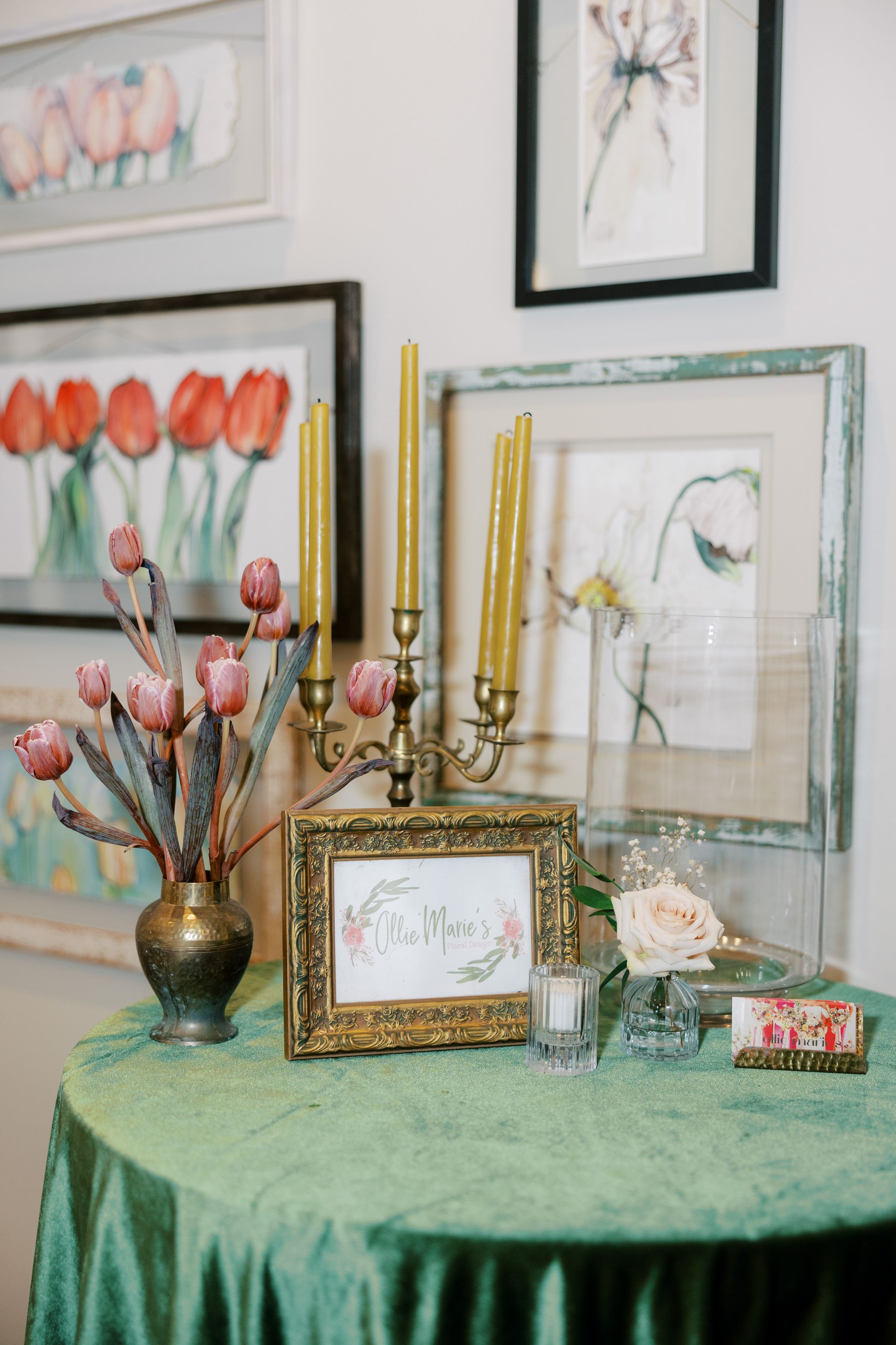 A table with floral arrangements, candles, and framed artwork against a wall decorated with framed paintings.