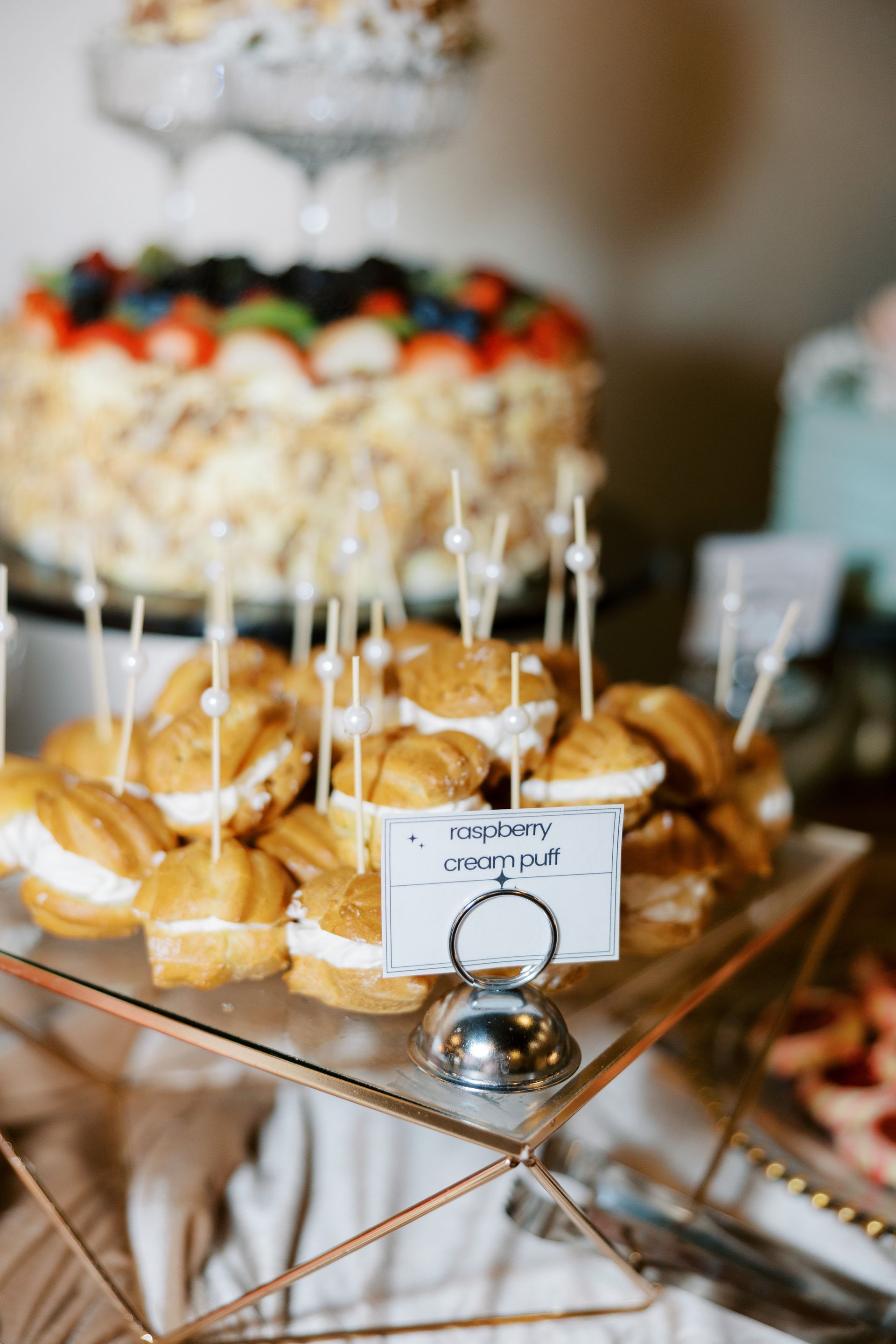 Display of cream puffs with pearl toothpicks, on a glass tray, with a cake in the background.