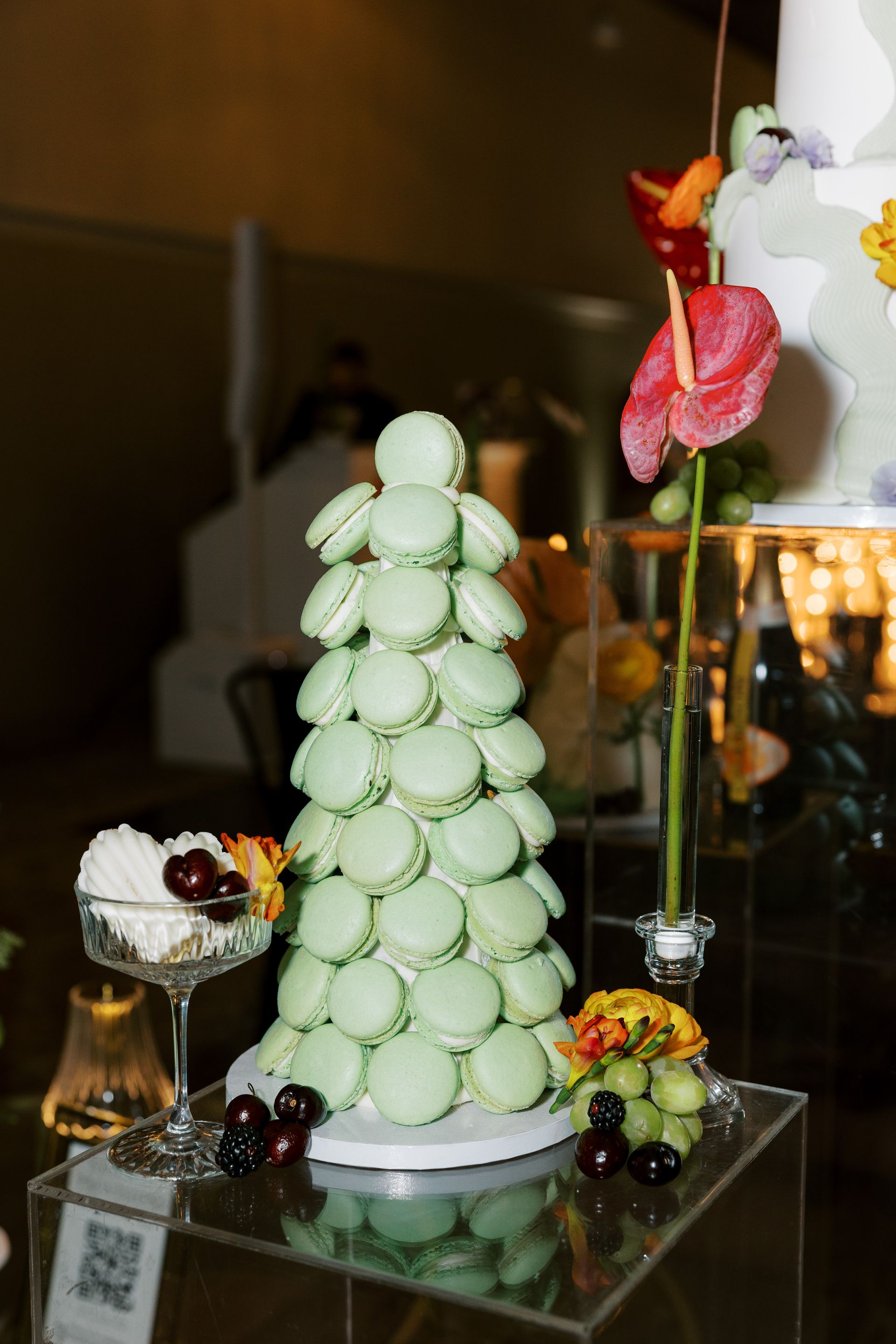 Green macaron tower on a white plate, with desserts and flowers on a clear table.