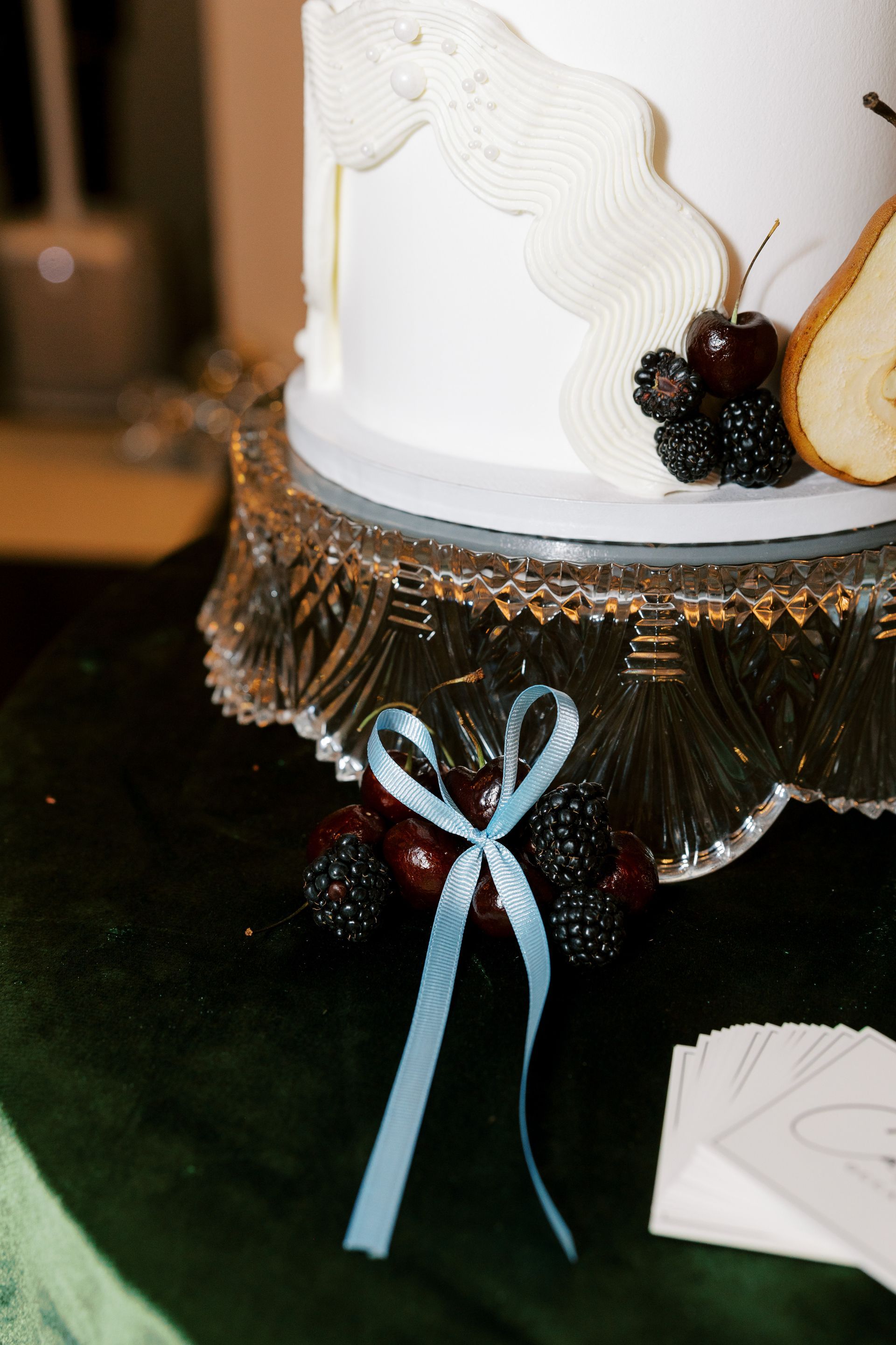 White cake with cream frosting and fruit decor, set on a glass cake stand, light blue ribbon accent.