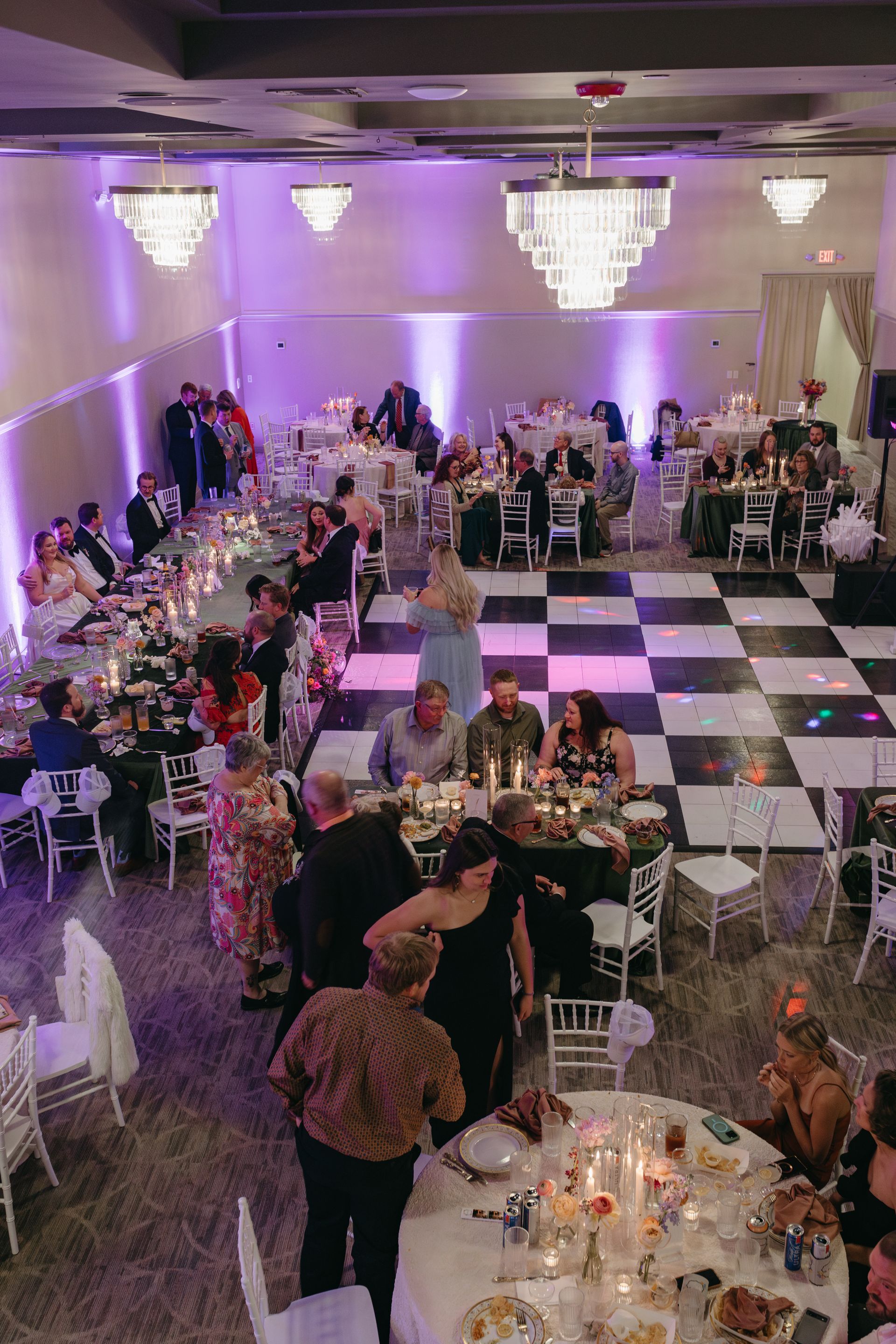 Wedding reception: Guests seated at tables in a large ballroom with a checkered dance floor and chandeliers.