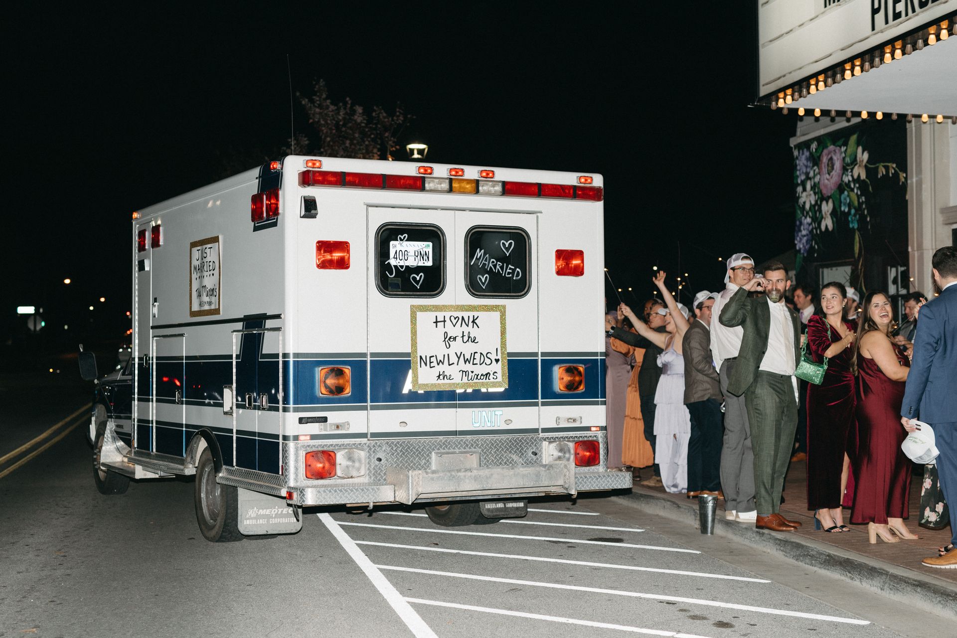 Ambulance parked at night near a theater; wedding guests wave and cheer.
