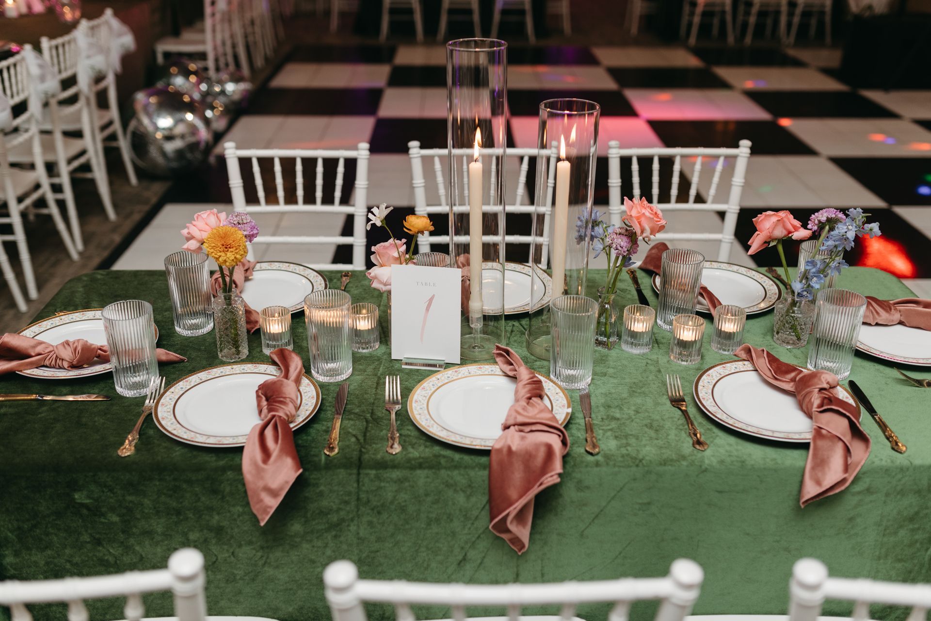 Elegant table setting with green tablecloth, gold-rimmed plates, pink napkins, candles, and flowers; checkered floor.