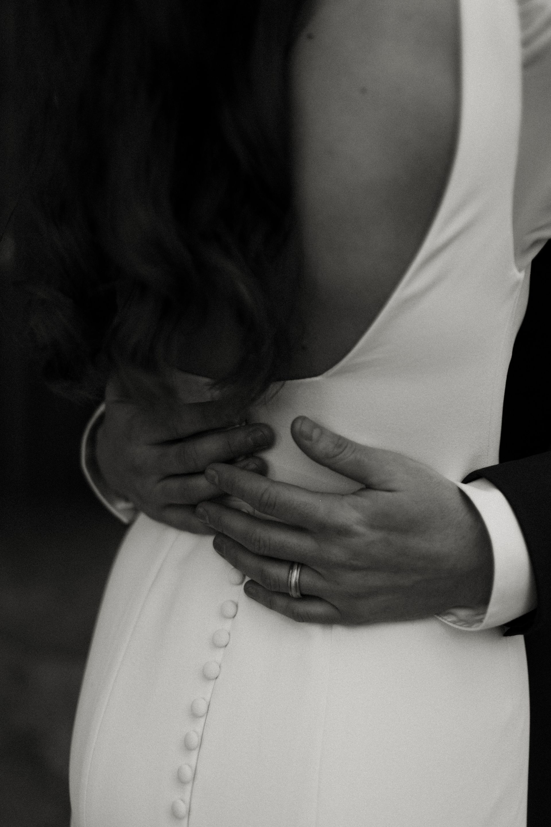 Hands of a person in a suit embrace a person in a white gown from behind; a ring is visible.