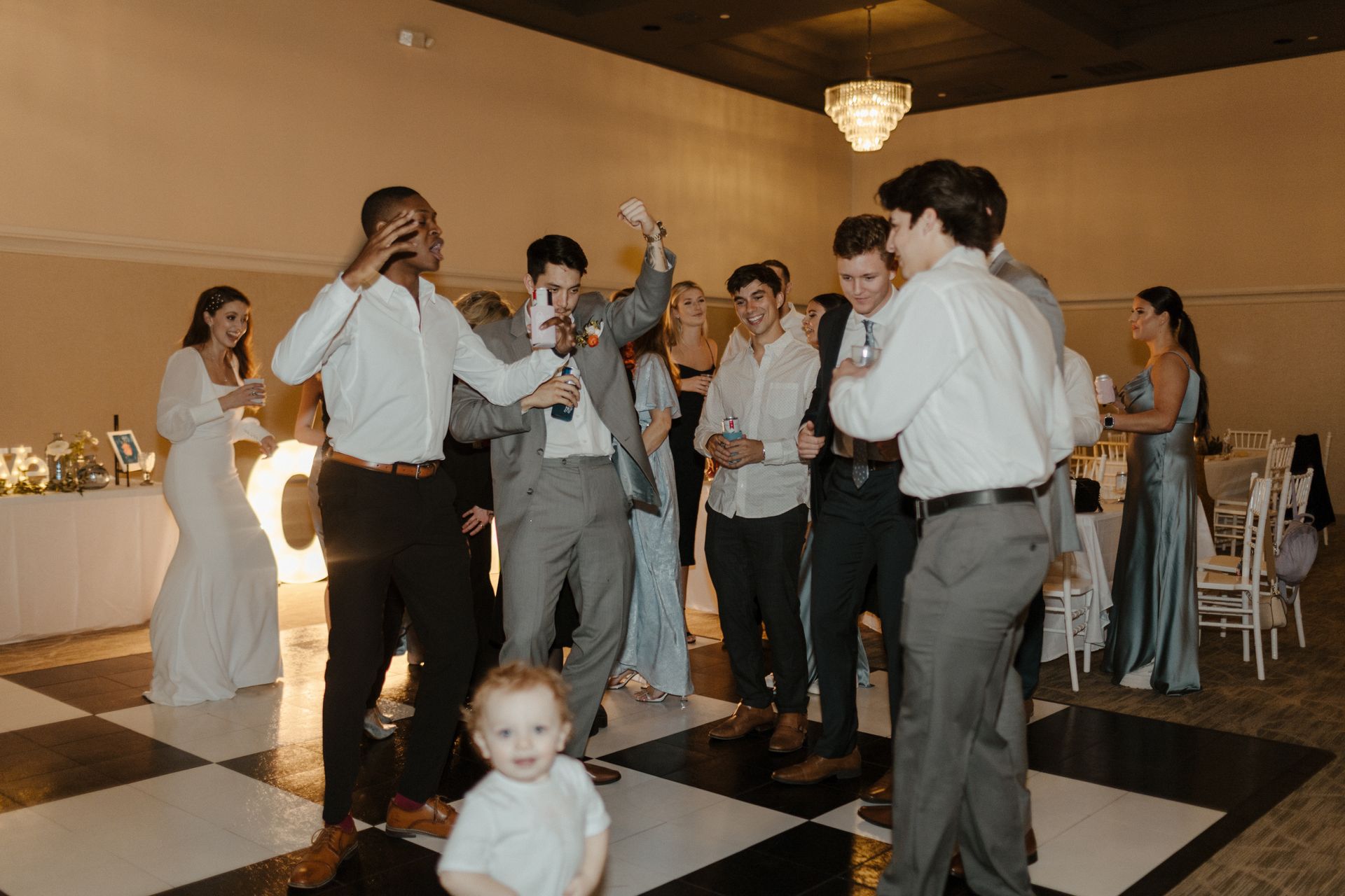 Wedding reception: Guests dance on a checkered floor. One holds a microphone, a child looks at the camera.