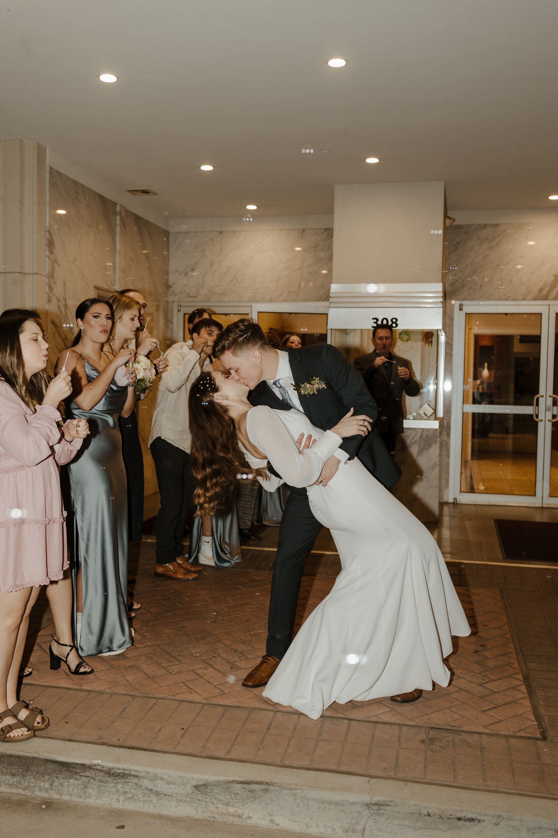 Bride and groom kissing outside. Guests with bubbles and confetti. Building entrance at night.