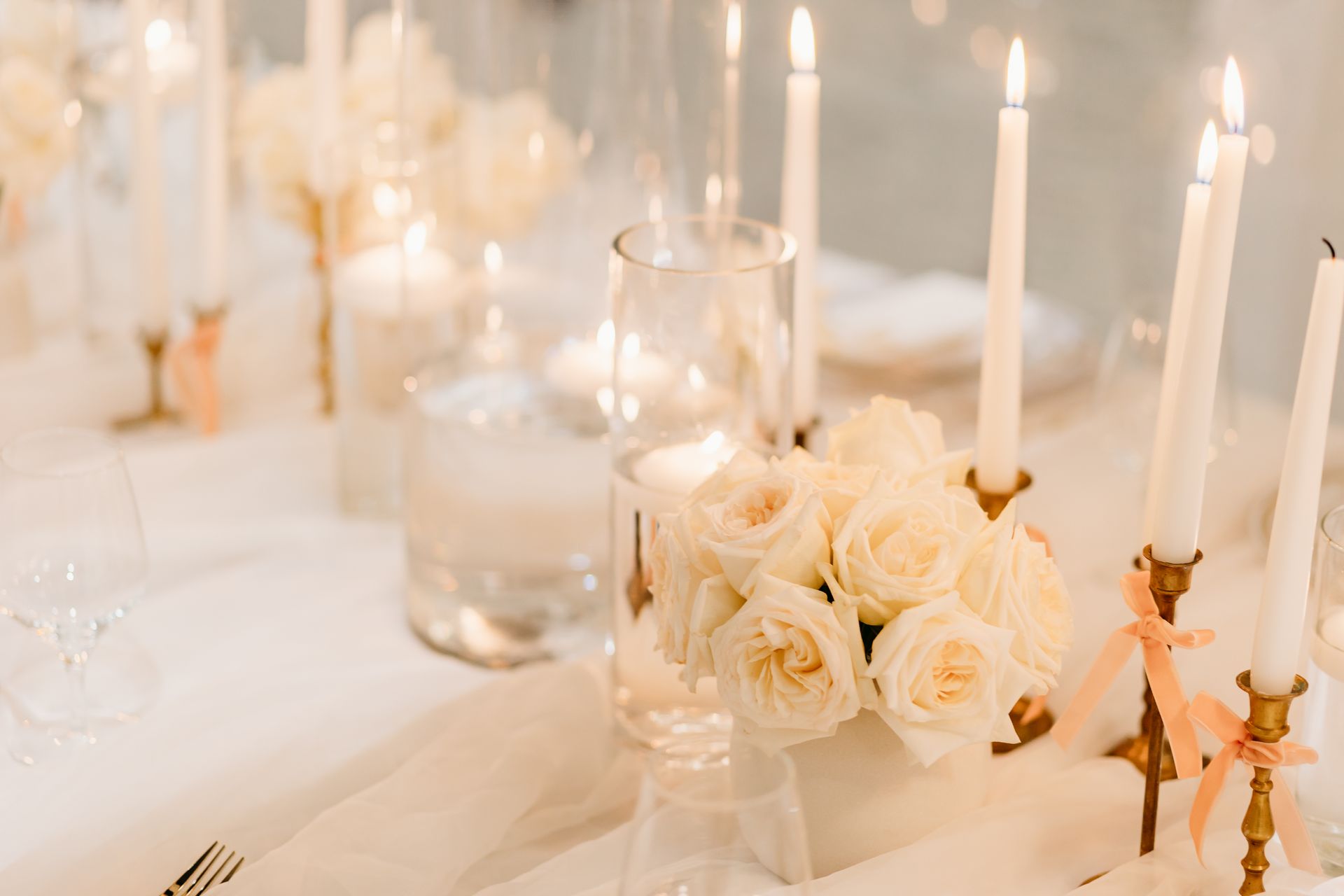 Elegant table setting with white candles, roses, and glassware, softly lit.