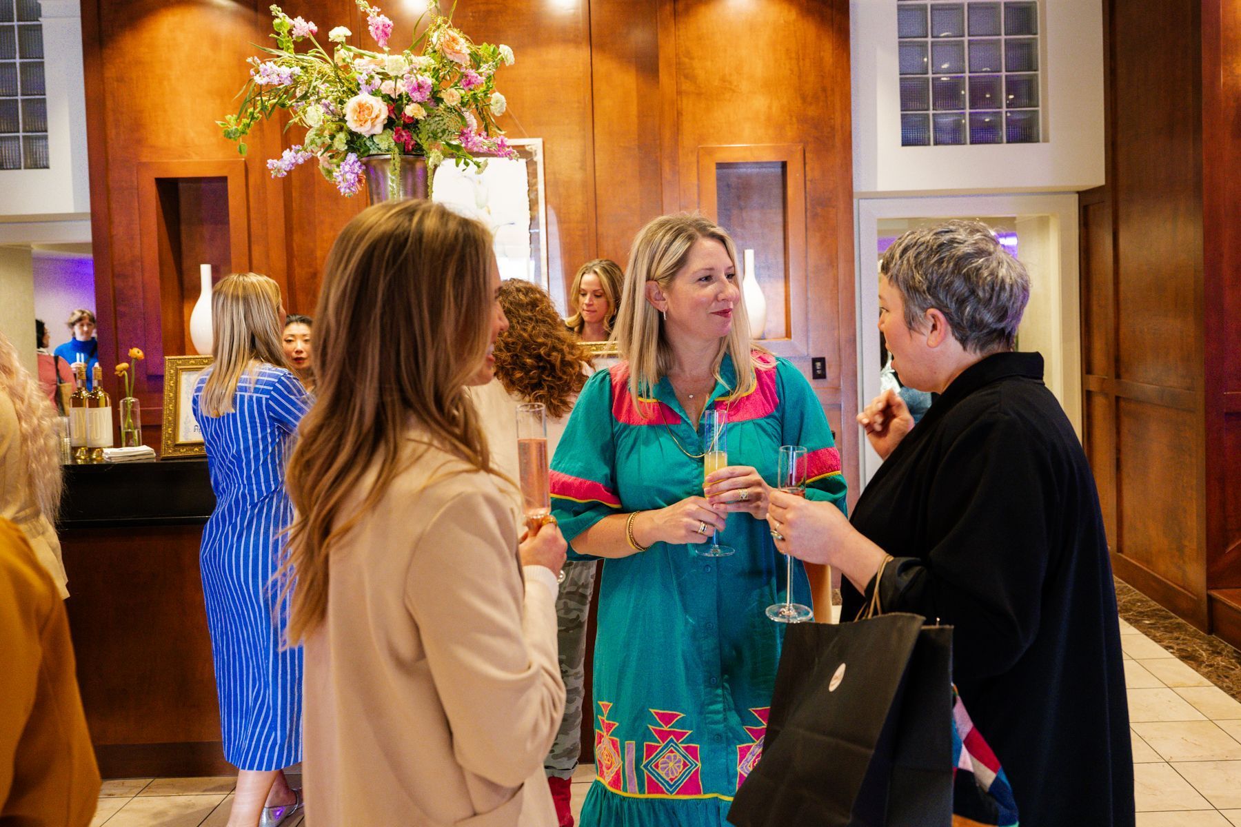 People socializing indoors with drinks, flower arrangement, wooden walls, light-colored stone floor.