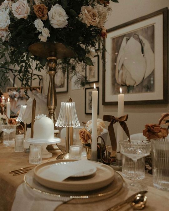 Elegant dining table set for a special occasion. Floral centerpiece, candles, framed artwork, and place settings with gold accents.