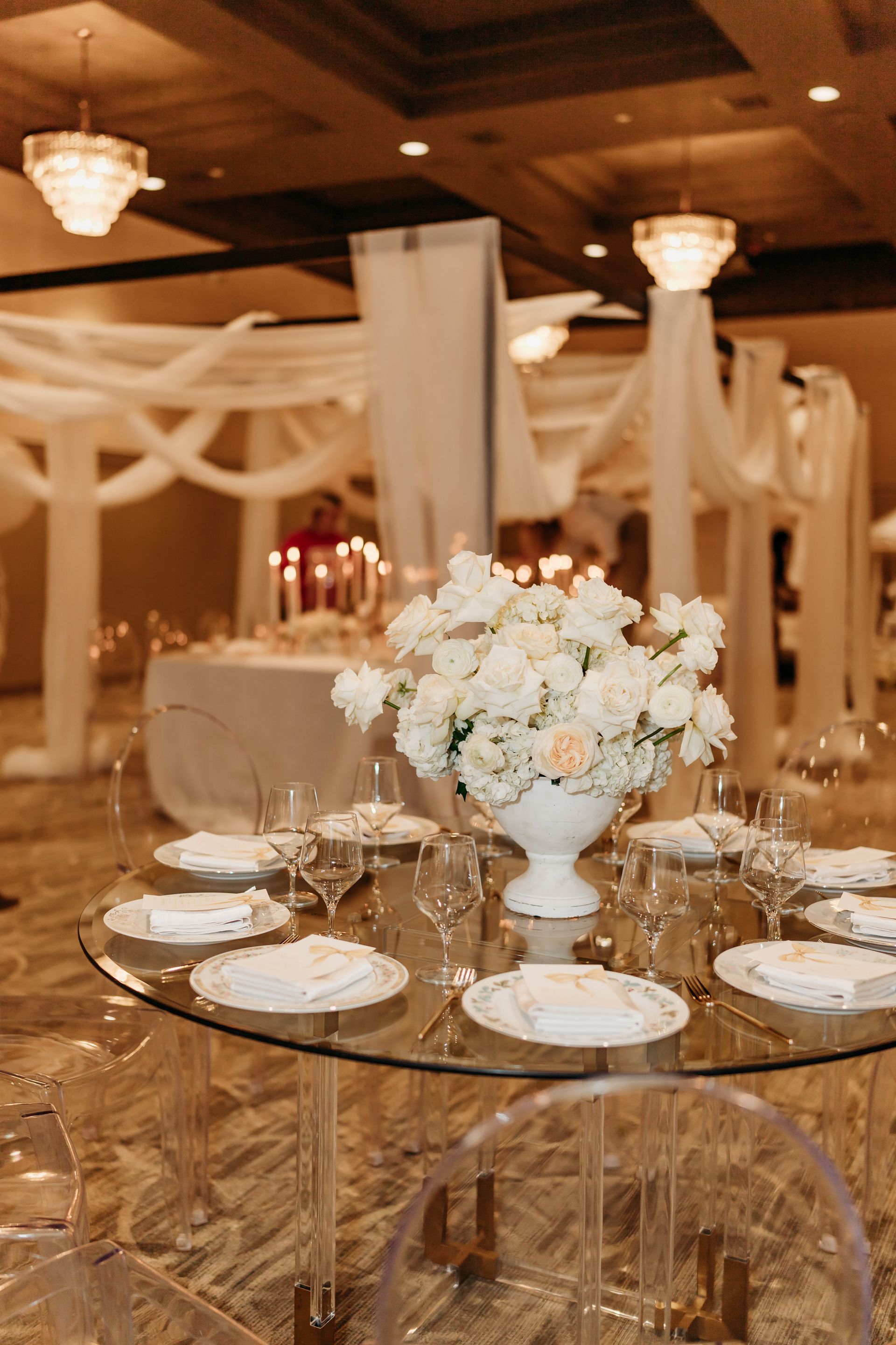 Round table with floral centerpiece, set for a formal event. Sheer white drapery, and crystal chandeliers.