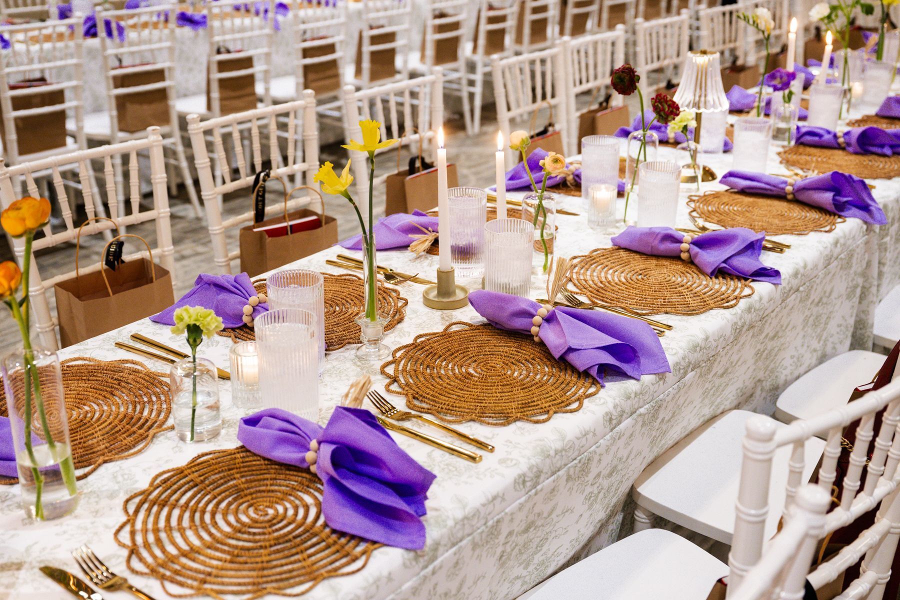 Long dining table set for event with gold chargers, purple napkins, white chairs.