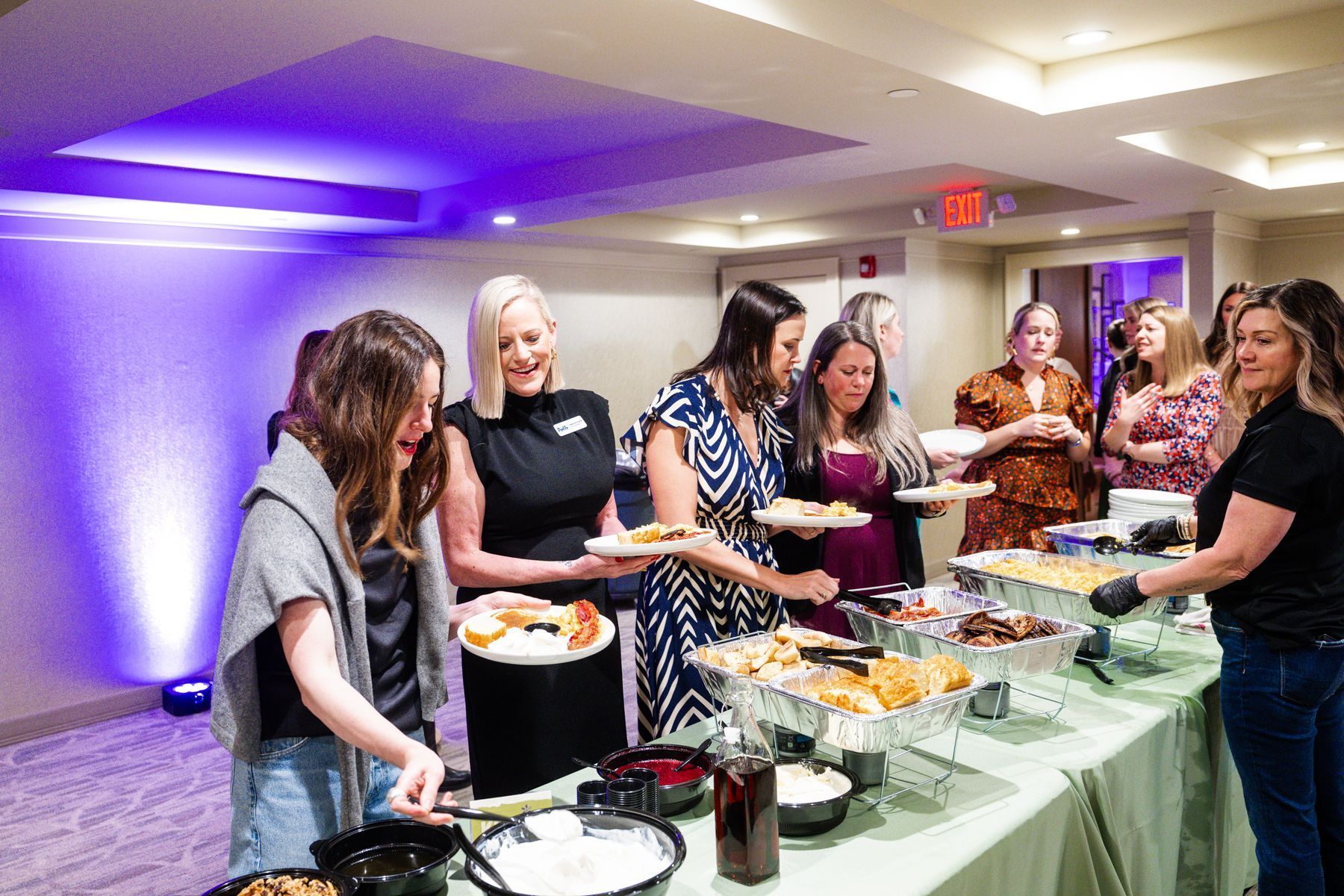 People at a buffet. Women gather around a table with food, taking plates of food. Green table cloth. Purple lighting.
